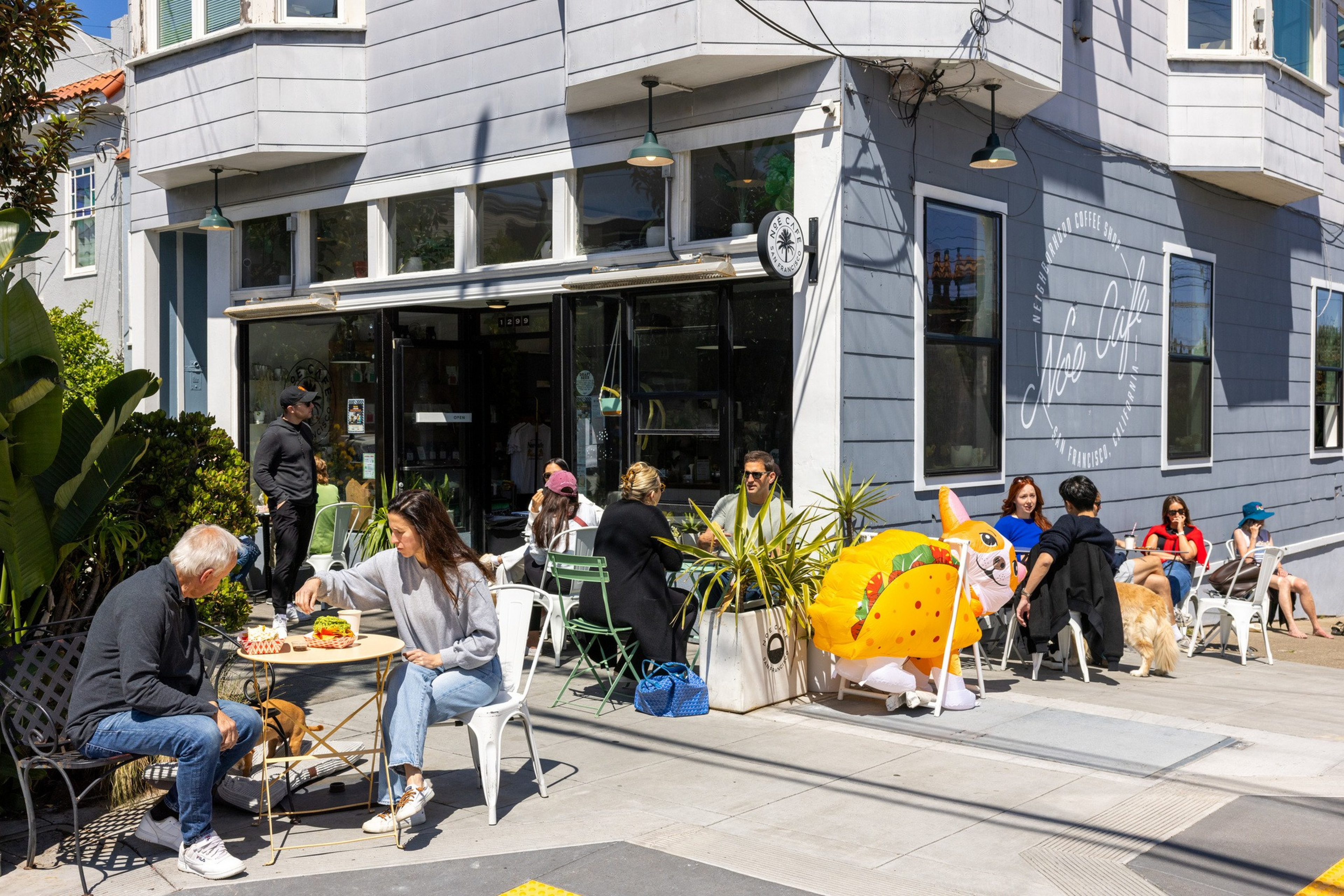 People sit and eat outdoors at a corner café on a sunny day, with a person in a colorful taco costume and a golden retriever nearby.