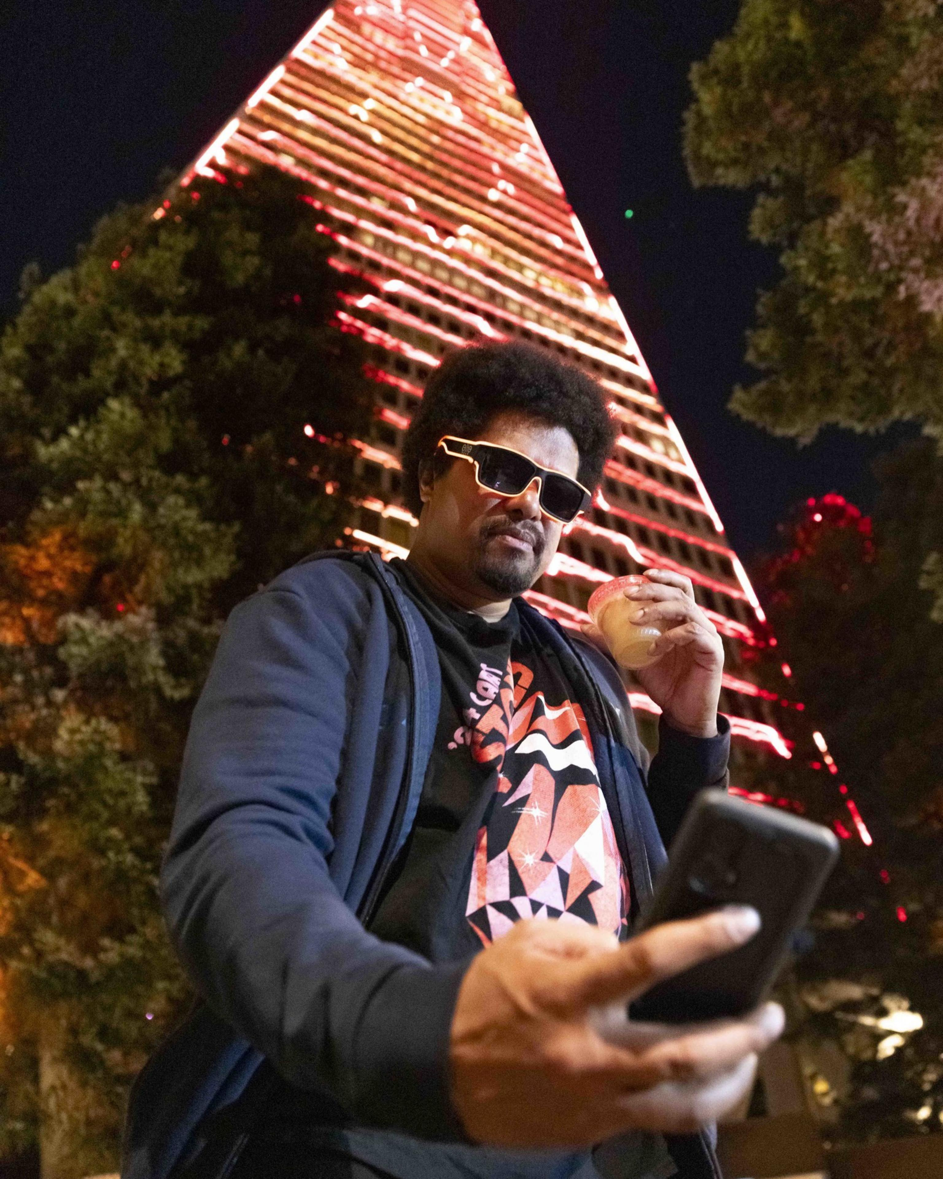A man wearing sunglasses holds a drink in one hand and a phone in the other, posing in front of a tall, illuminated, triangular building at night.