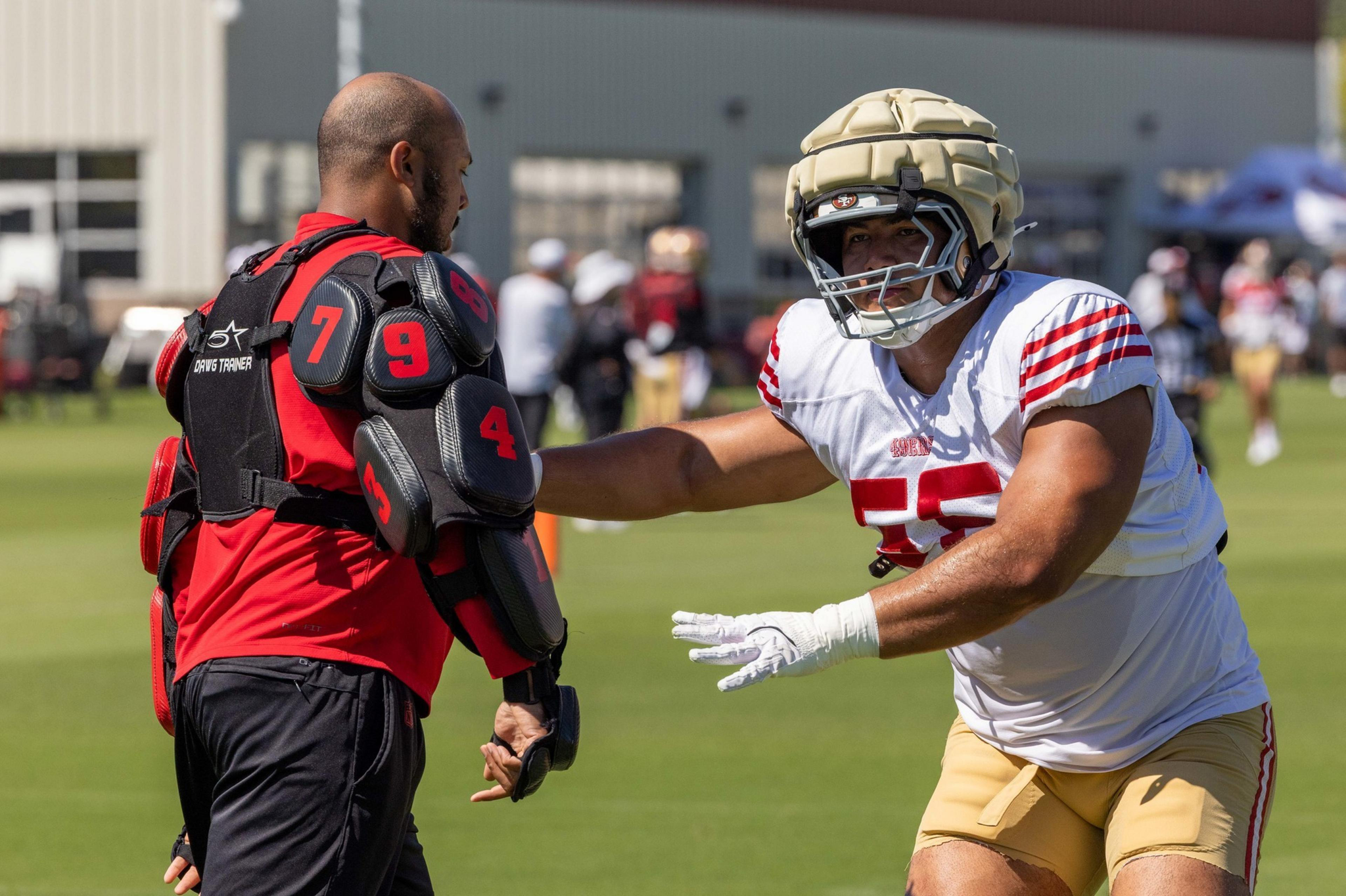 A football player in gold pants and white jersey with red stripes reaches toward a trainer in red and black padded gear on a grassy field.