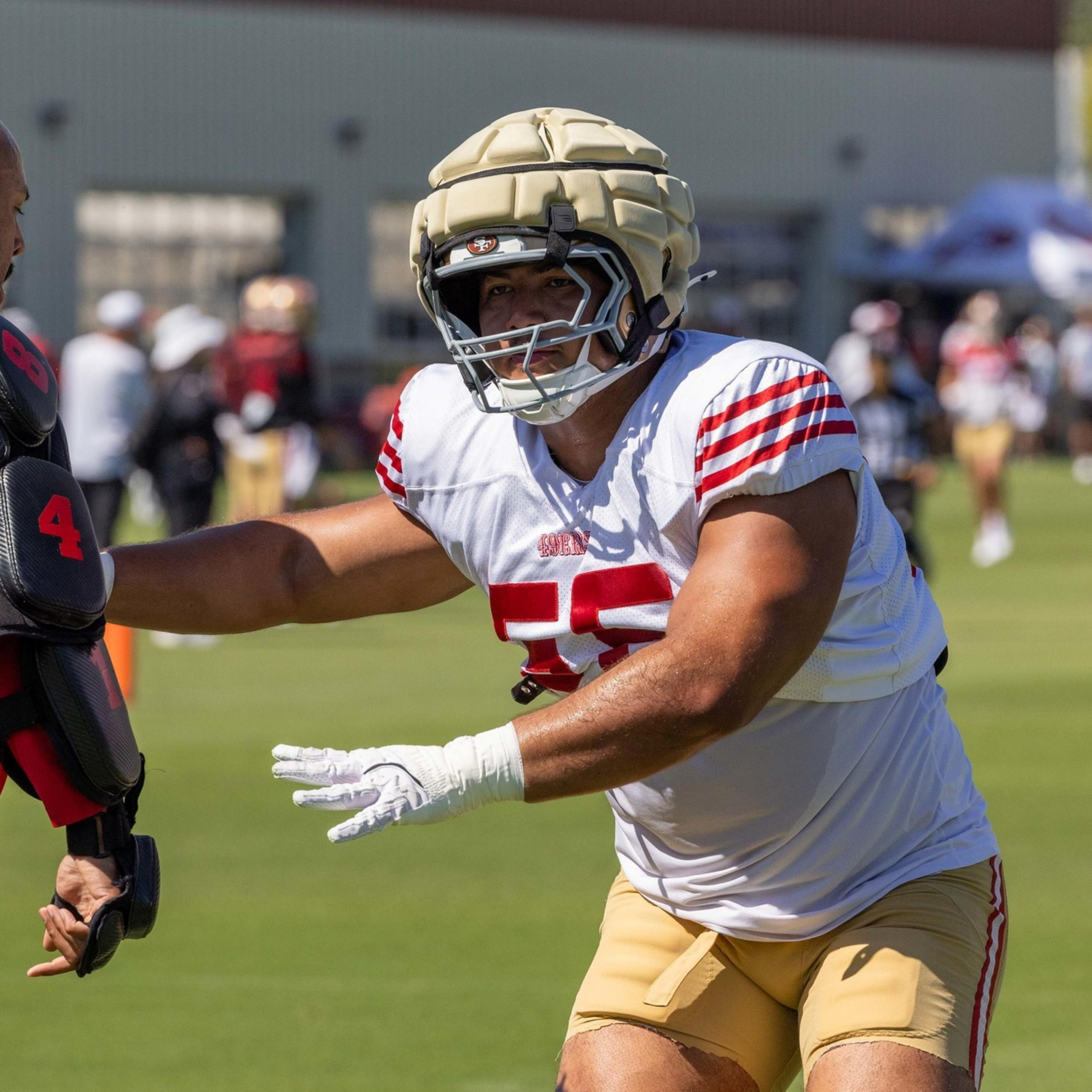 A football player in gold pants and white jersey with red stripes reaches toward a trainer in red and black padded gear on a grassy field.