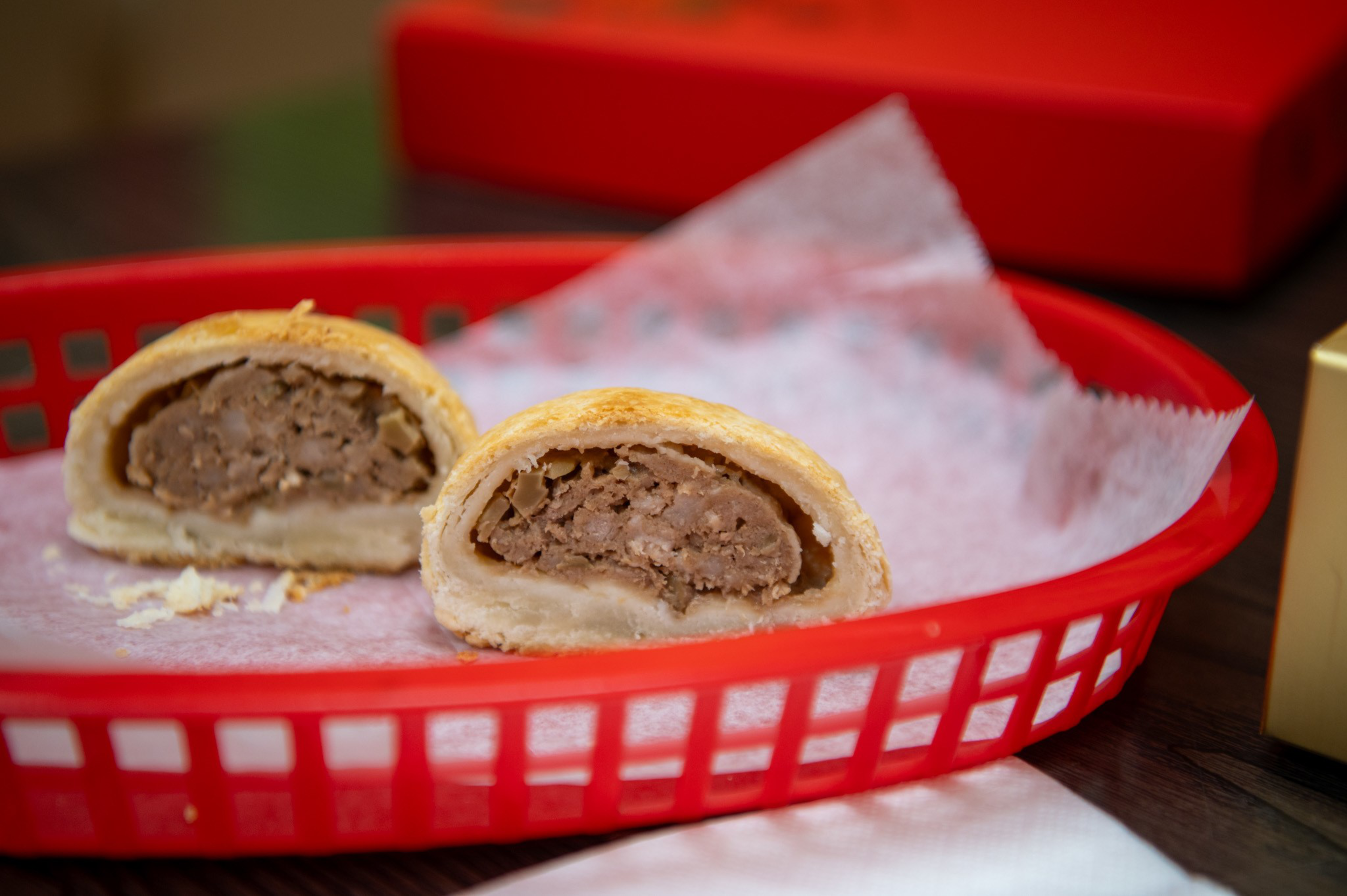 Two halves of a meat-filled pastry sit on white parchment paper inside a red plastic basket on a wooden table.