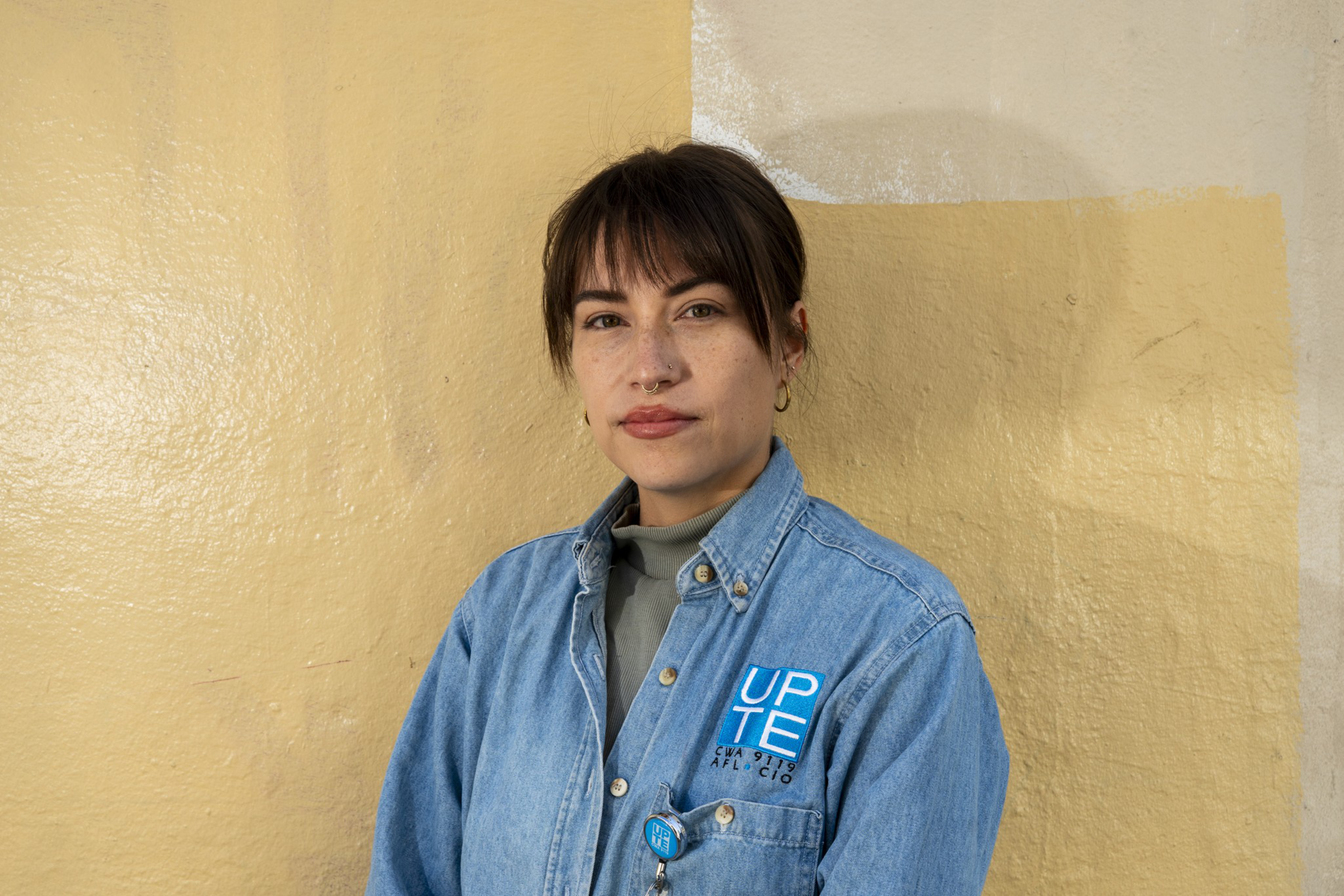 A woman with short dark hair and bangs wears a blue denim shirt with a UPTA logo, standing against a beige wall with a neutral expression.