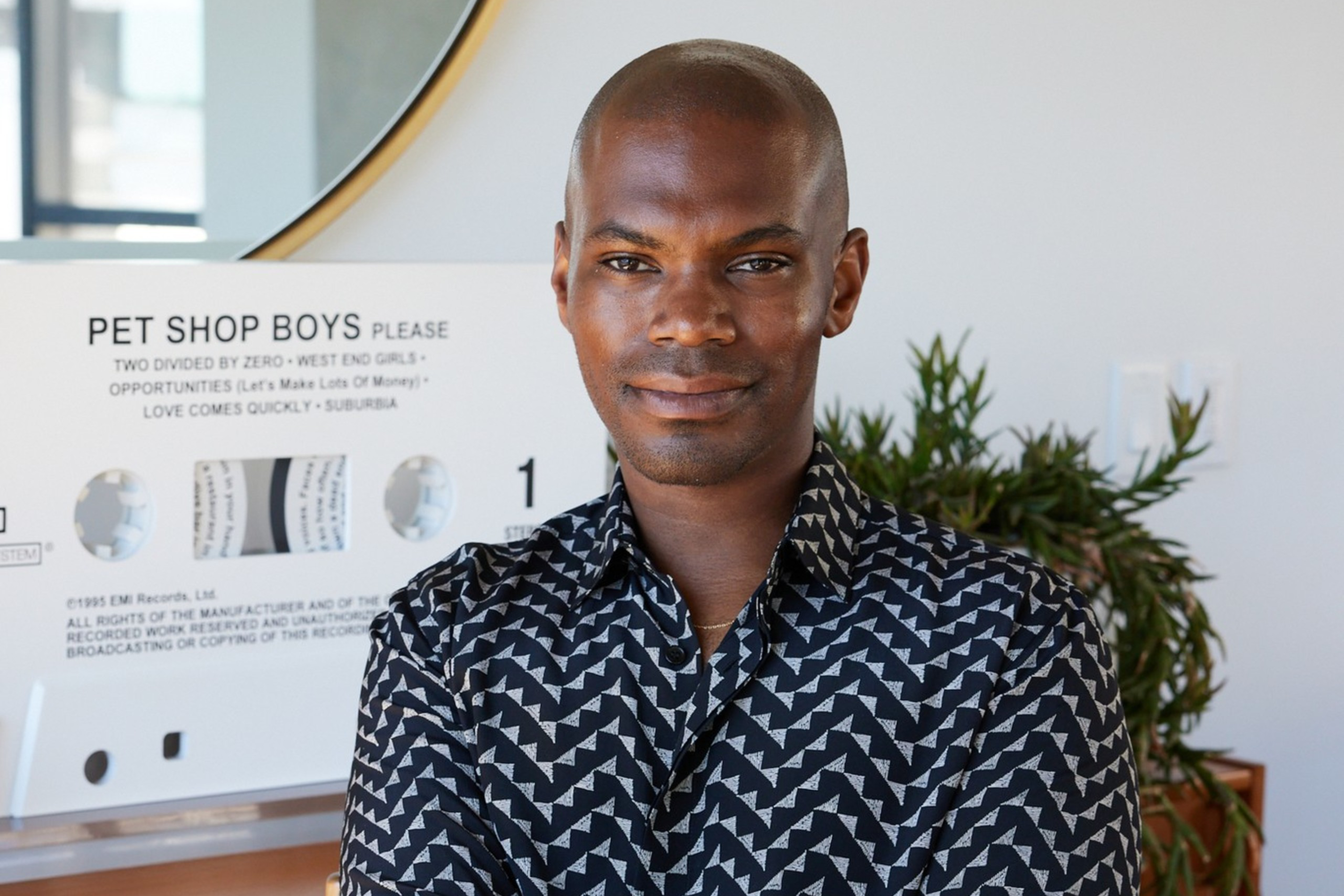 A person sits with an arm crossed, wearing a patterned shirt. Behind is a large circular mirror and a "Pet Shop Boys" sign. Thereâ€™s also a plant in the background.