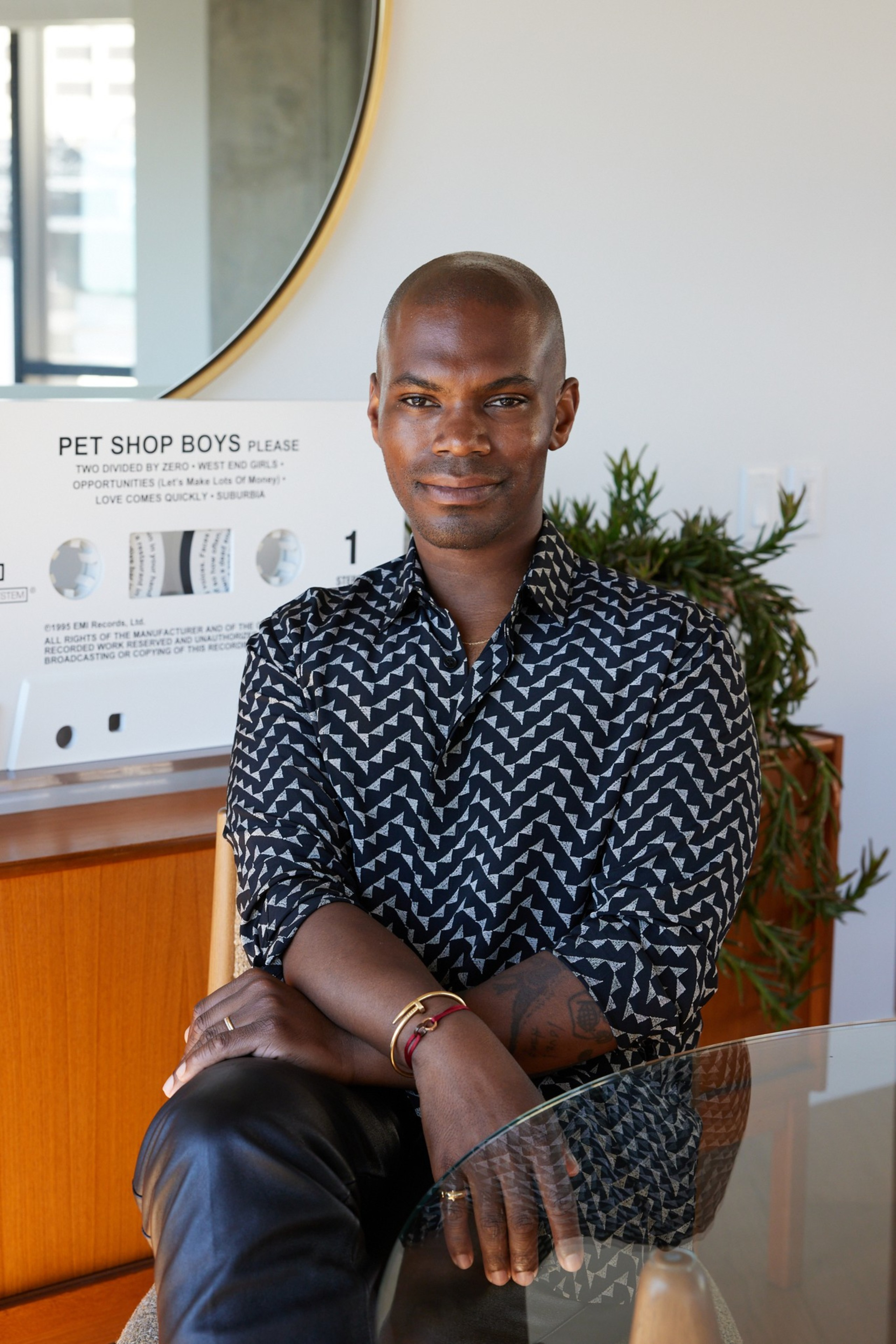 A person sits with an arm crossed, wearing a patterned shirt. Behind is a large circular mirror and a "Pet Shop Boys" sign. Thereâ€™s also a plant in the background.