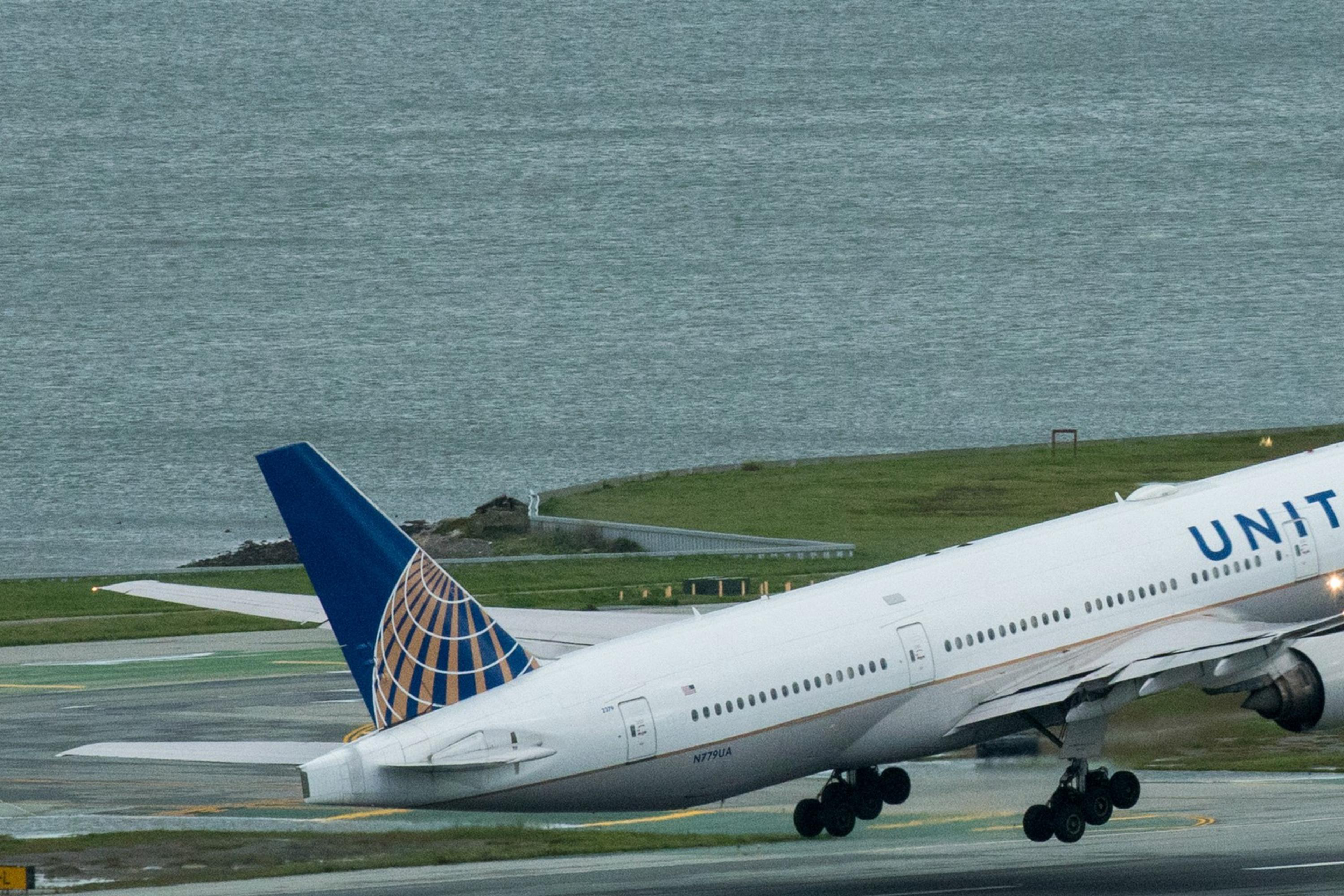 A white airplane with "United" on the side is taking off from an airport runway, with the ocean visible in the background on a cloudy day.