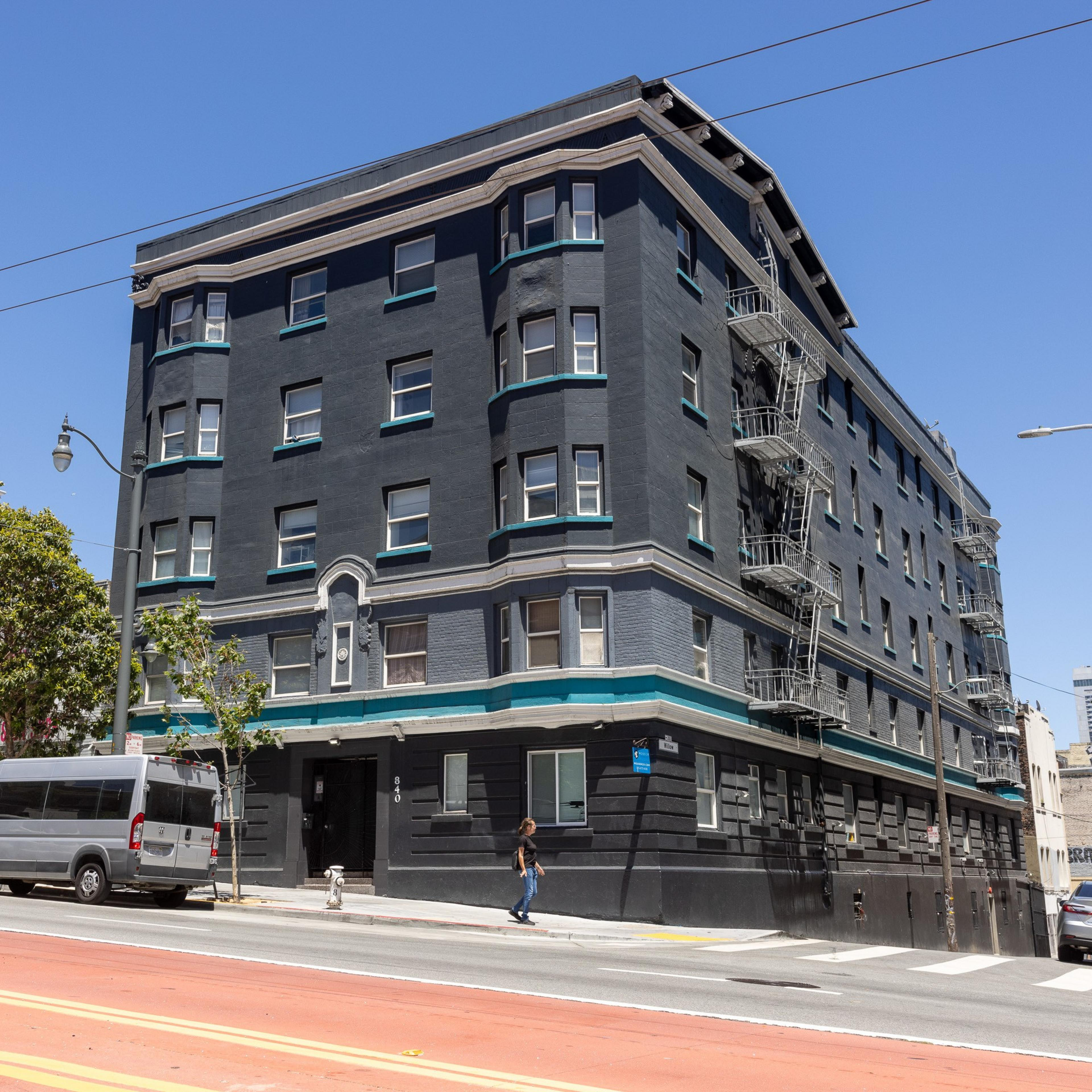 A dark gray, four-story building with teal trim and numerous windows stands on a street corner. A person walks on the sidewalk, and a white van is parked nearby.