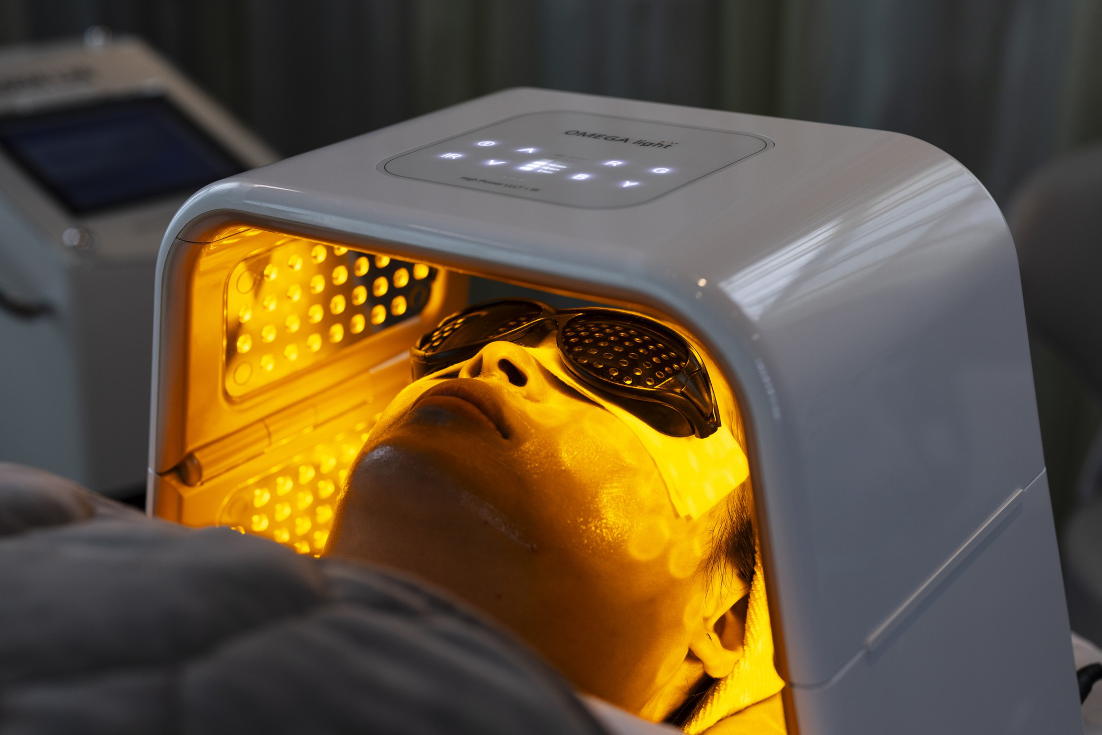 A person wearing protective goggles lies under a white LED light therapy device emitting orange light onto their face.