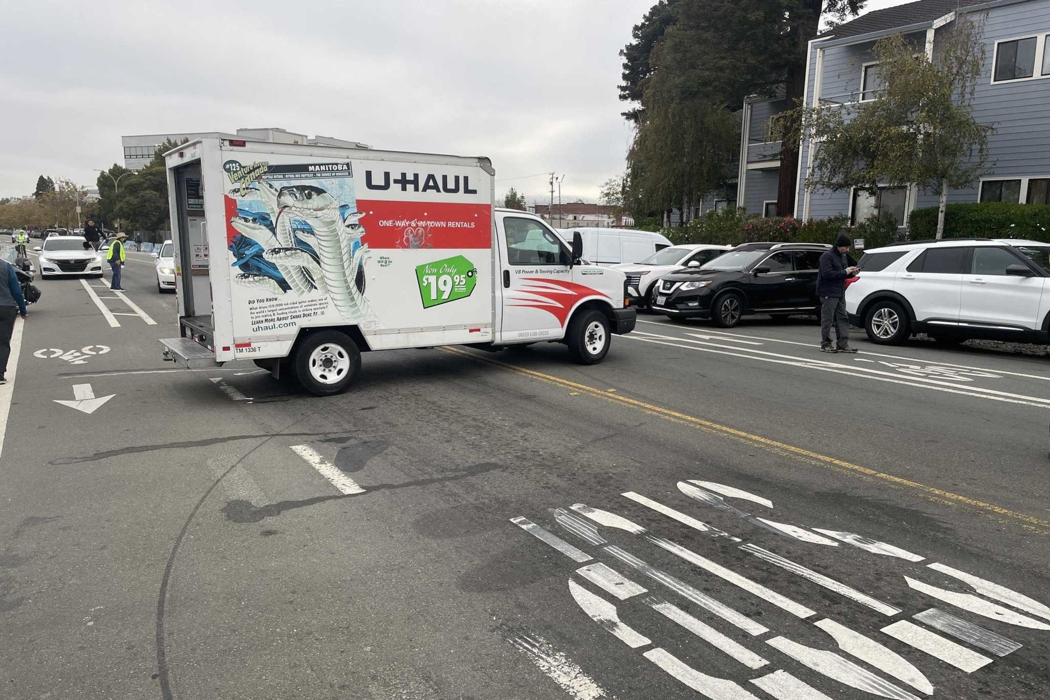 A U-Haul truck is blocking a bike lane and bus-only lane on a street with parked cars and a few people nearby.