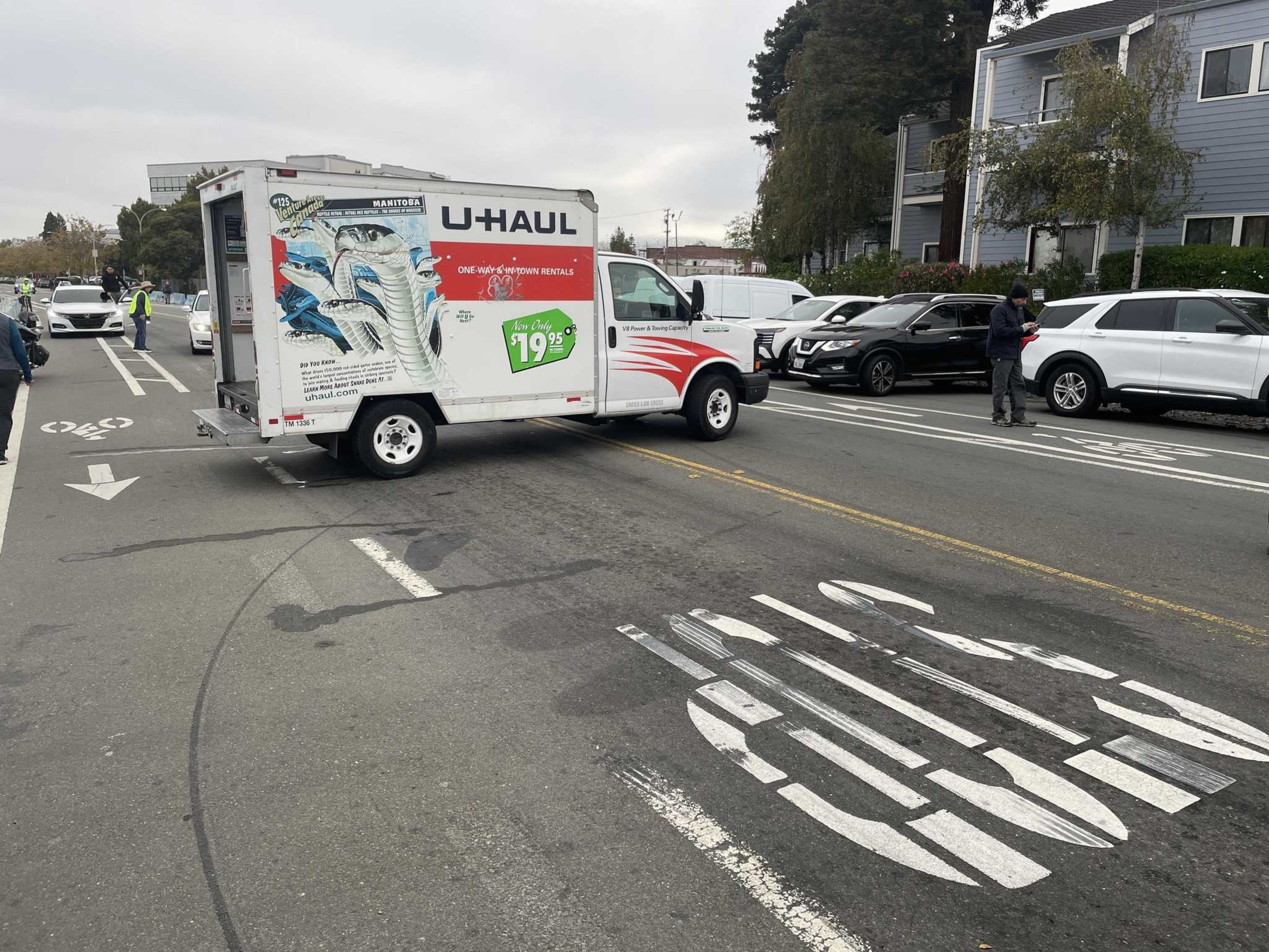 A U-Haul truck is blocking a bike lane and bus-only lane on a street with parked cars and a few people nearby.