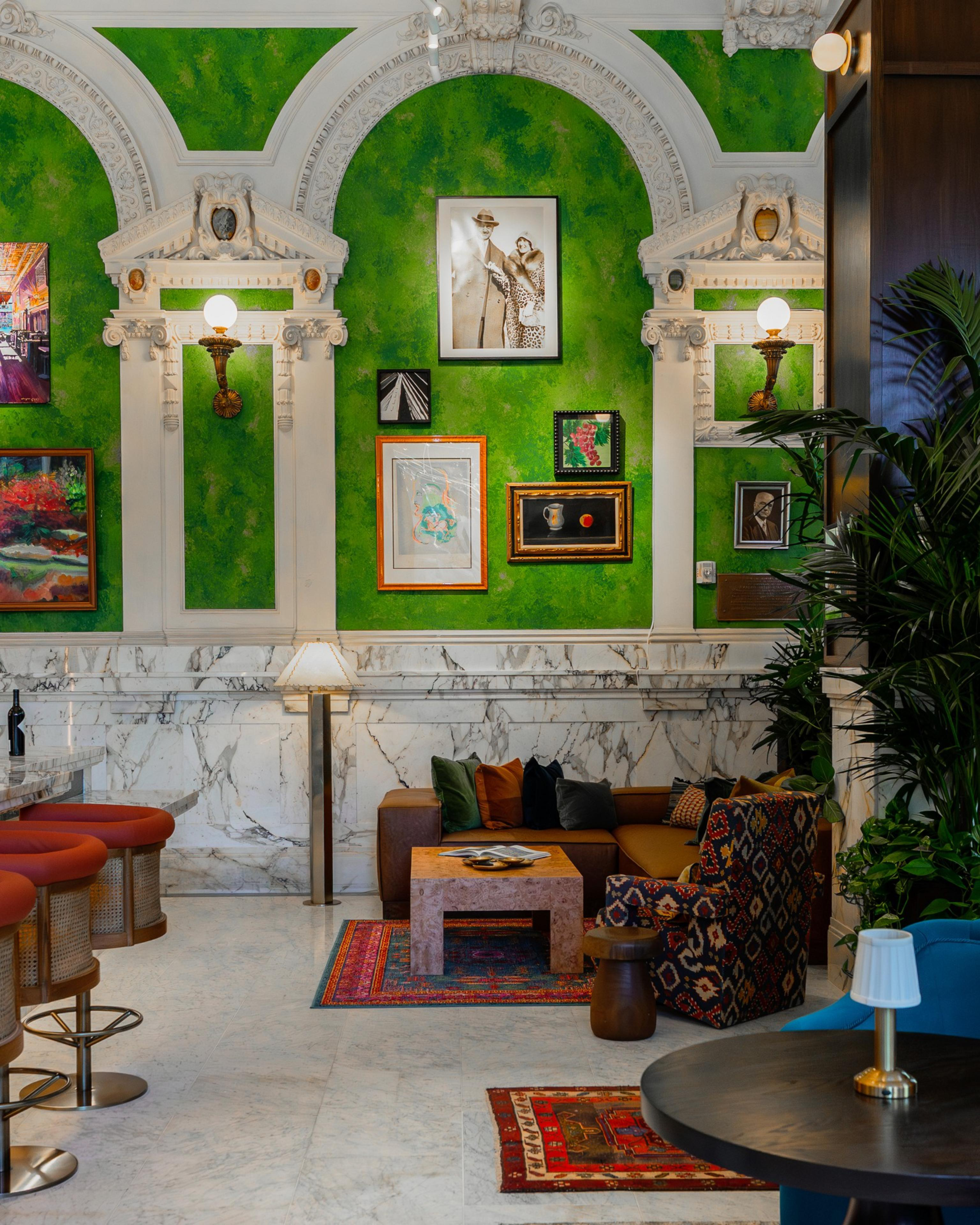 A vibrant green wall with white ornate arches and framed art above a brown couch with multicolored cushions, a patterned armchair, and rugs.