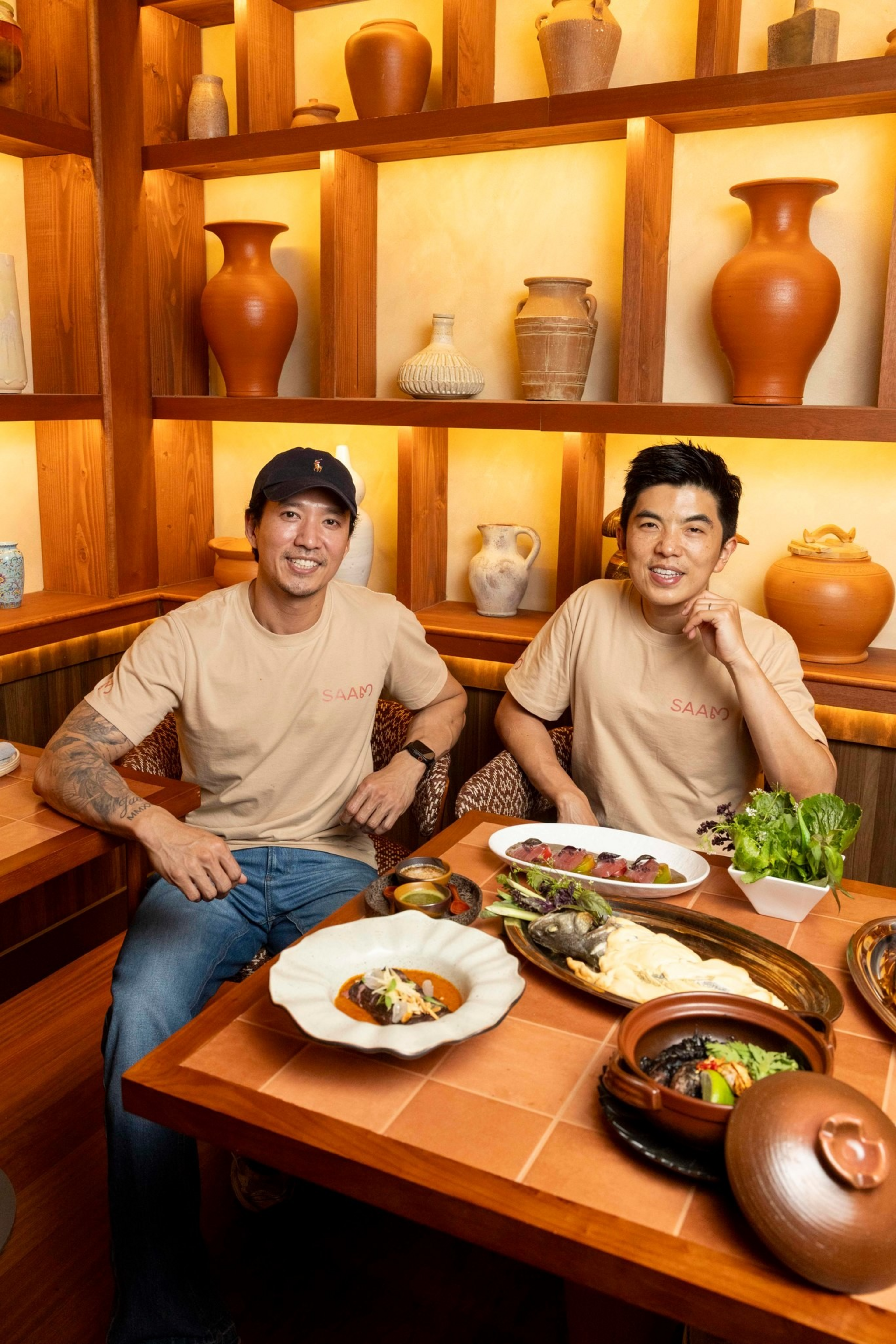 Two men wearing matching beige shirts sit at a wooden table with various plates of food, surrounded by shelves filled with clay pots.