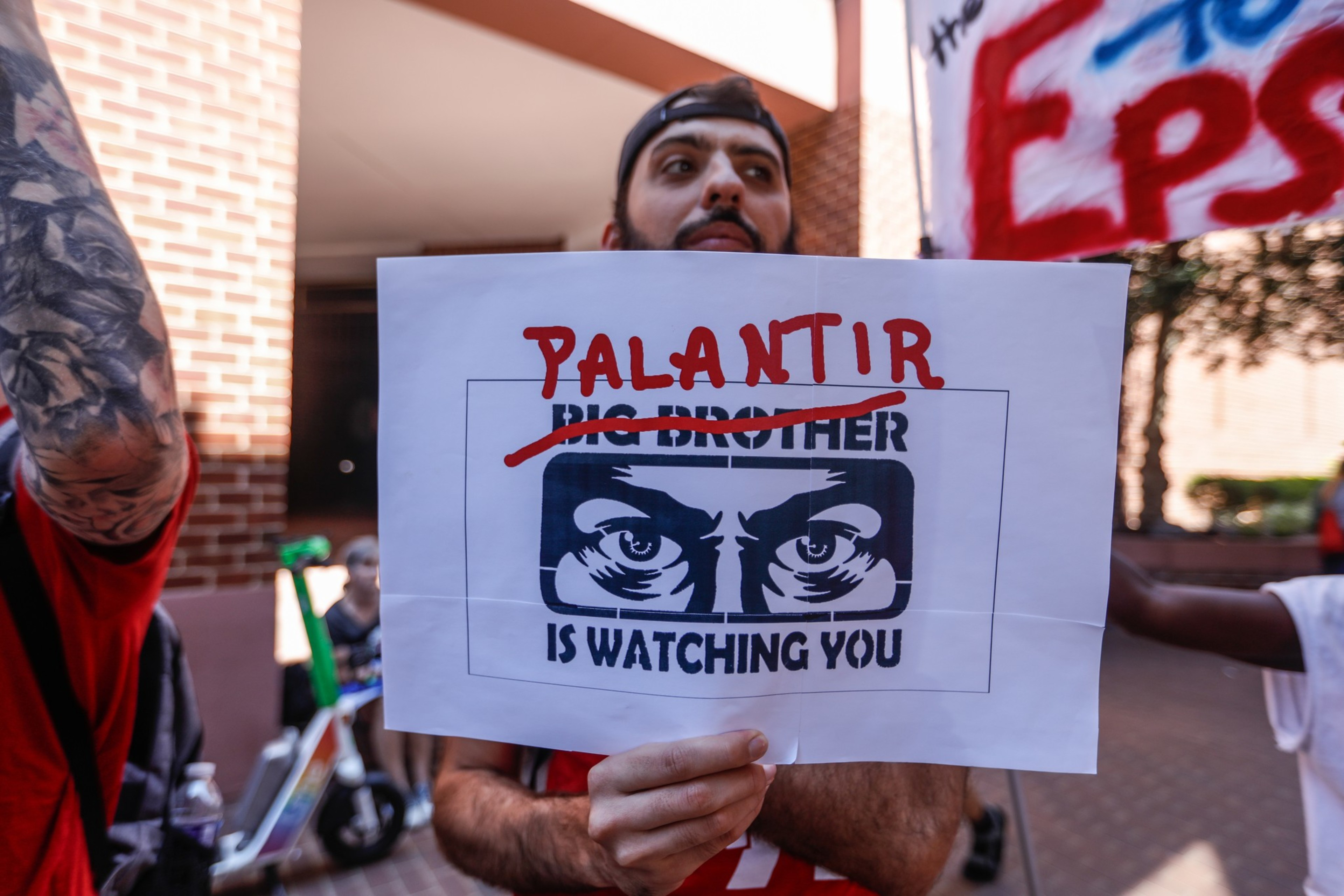 A person holds a sign with eyes and the words “BIG BROTHER IS WATCHING YOU,” with “BIG BROTHER” crossed out and “PALANTIR” written above in red.