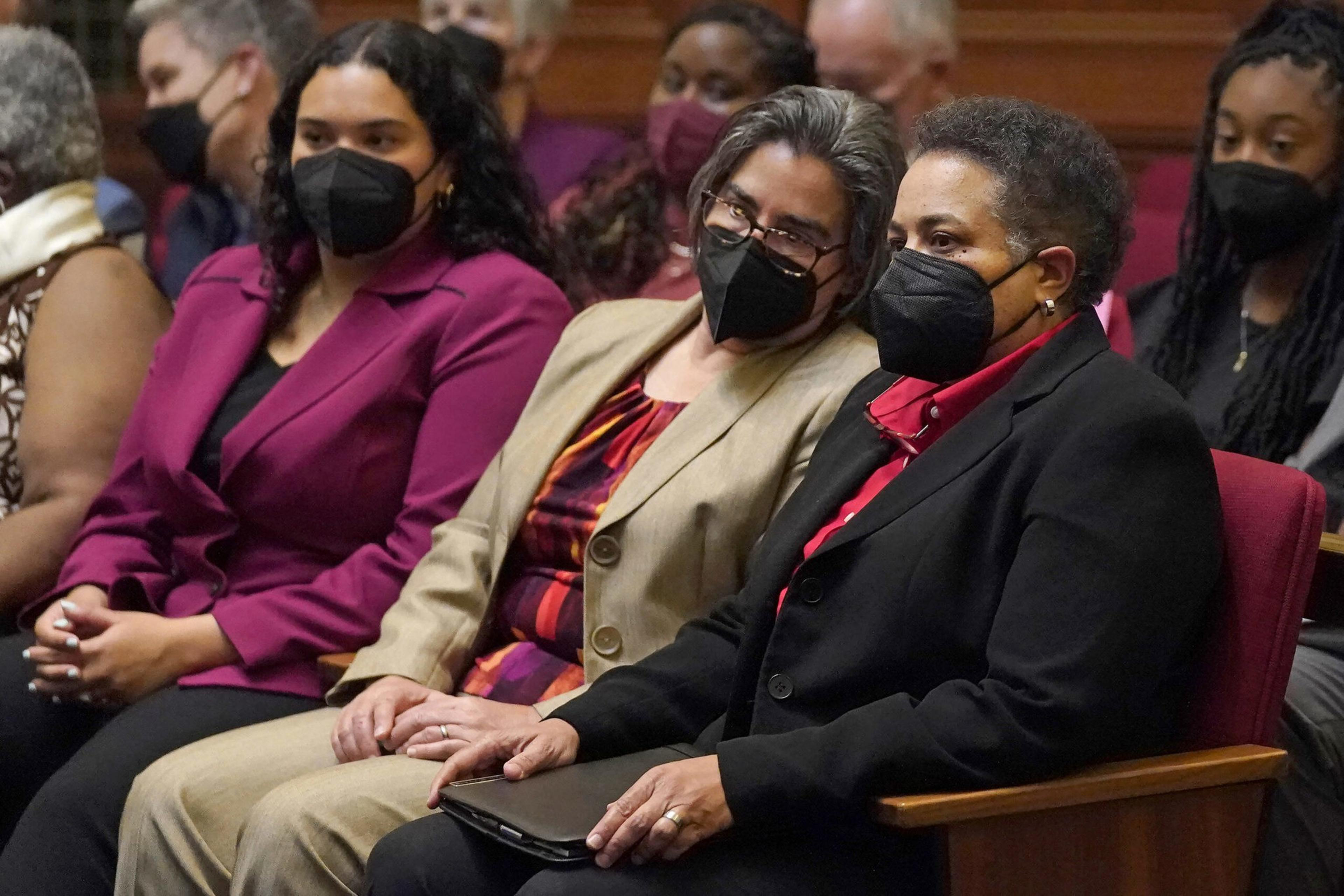 A group of people sit closely together wearing face masks, dressed in formal and business attire, appearing attentive and engaged.