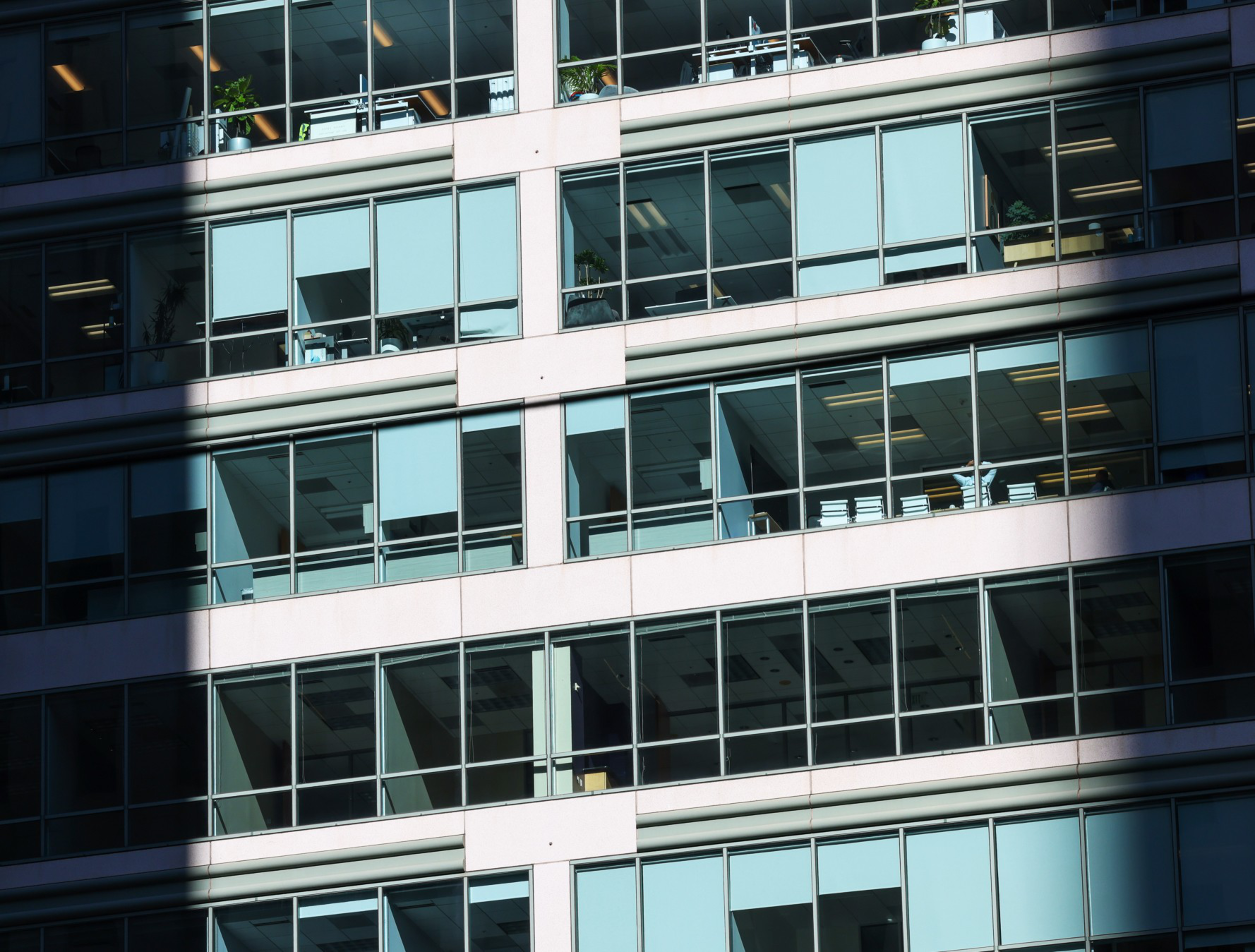 Glass windows of an office building reflect daylight, revealing mostly empty rooms with desks and some plants on the upper floor.