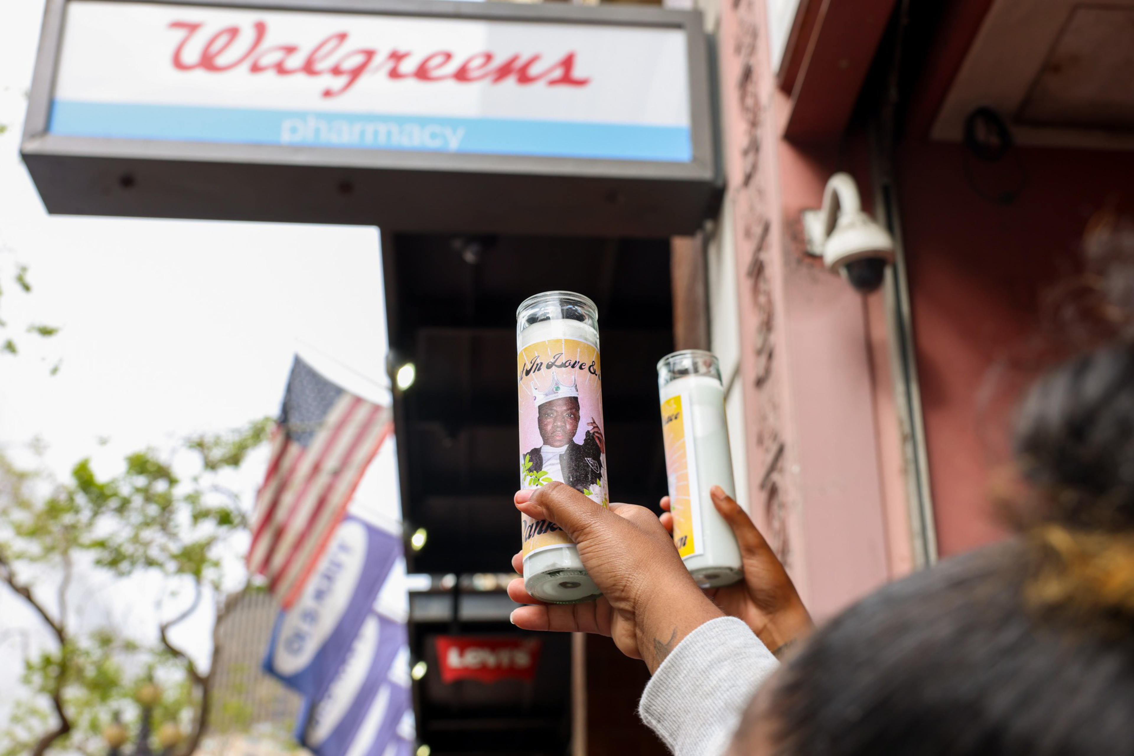 Two hands hold memorial candles, one with a photo of a man, raised near a Walgreens pharmacy sign and an American flag.