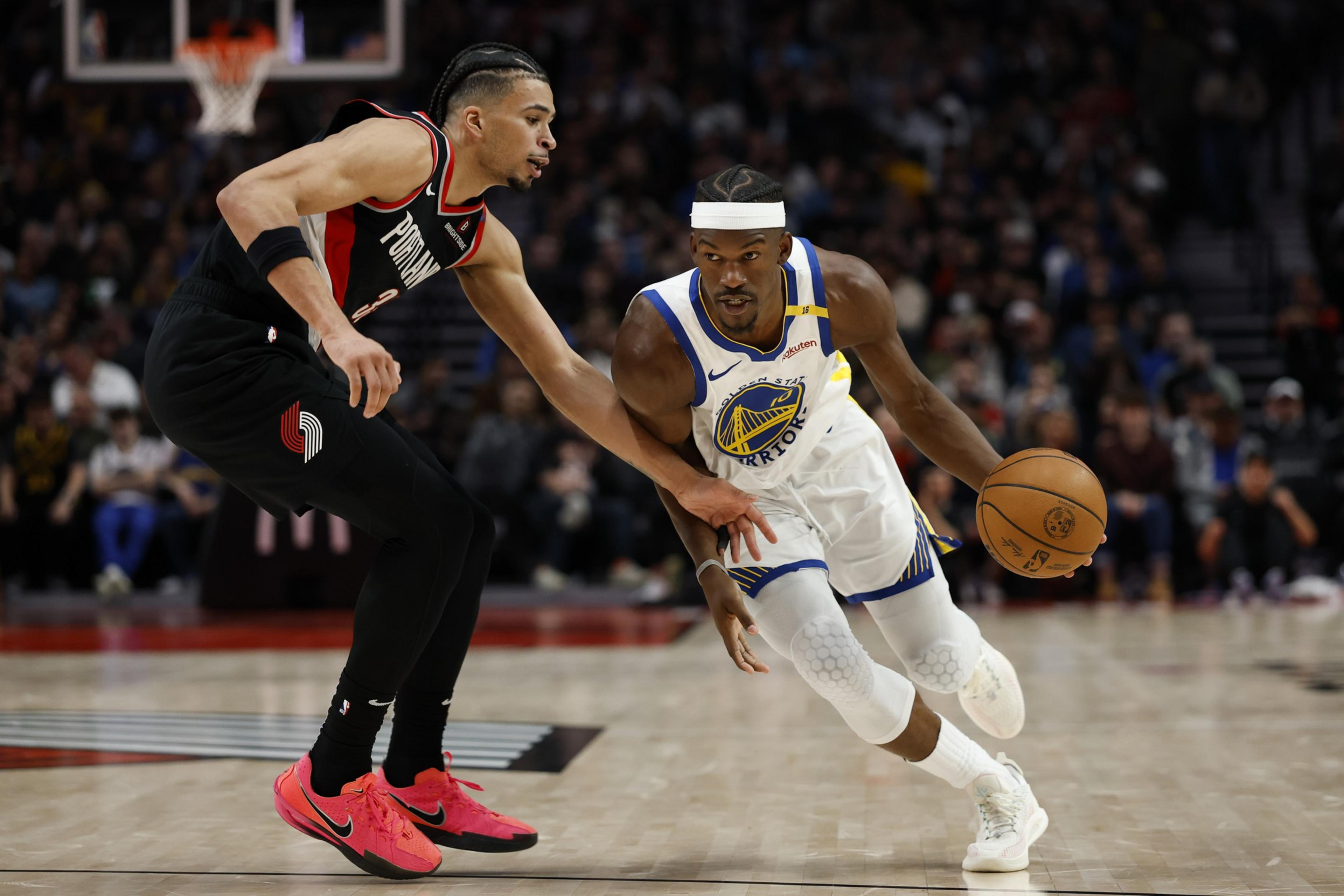 One basketball player in a white and blue uniform dribbles past another in a black uniform on the court, in front of a blurred crowd.