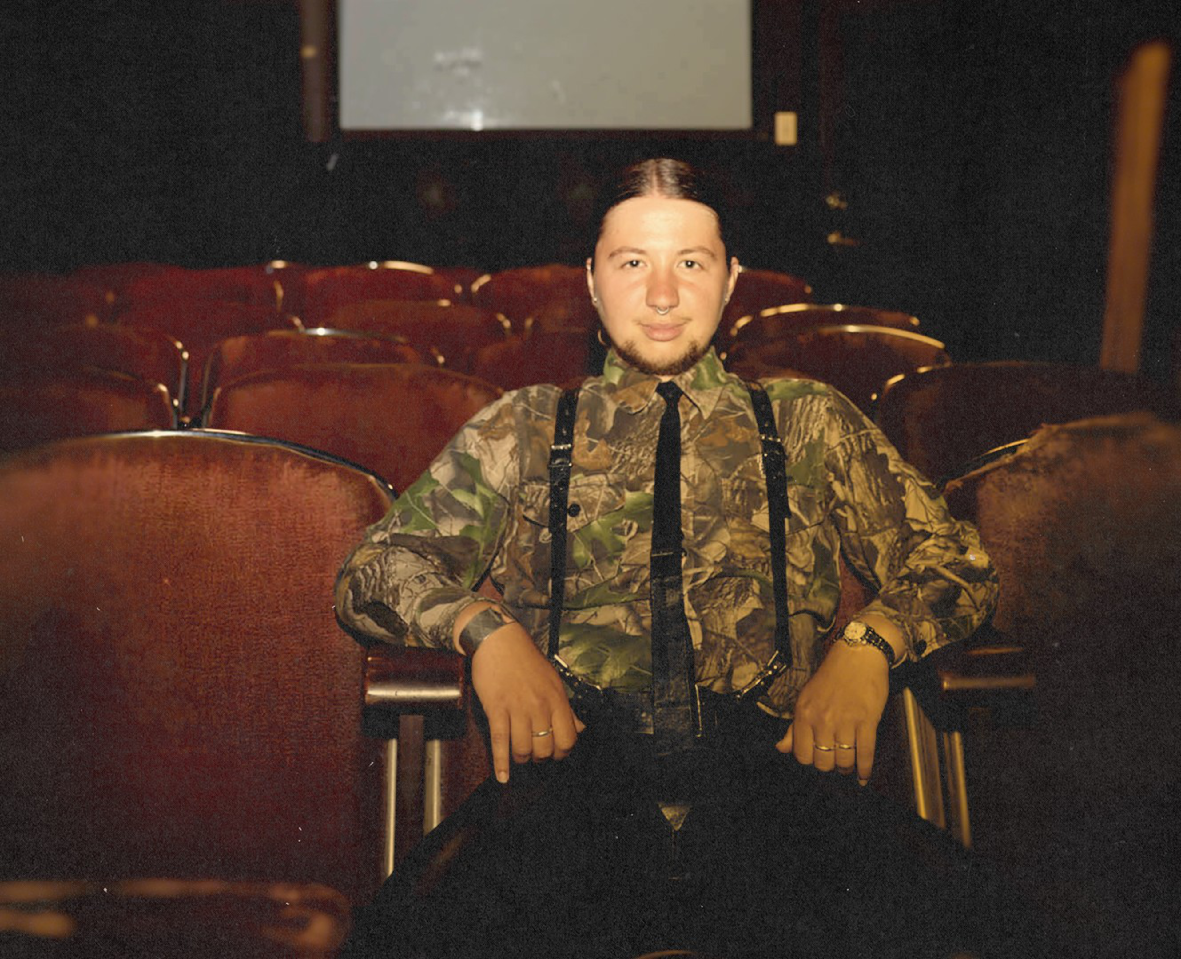 A person with a camo shirt, black suspenders, and a black tie sits relaxed in an empty auditorium with brown cushioned seats.