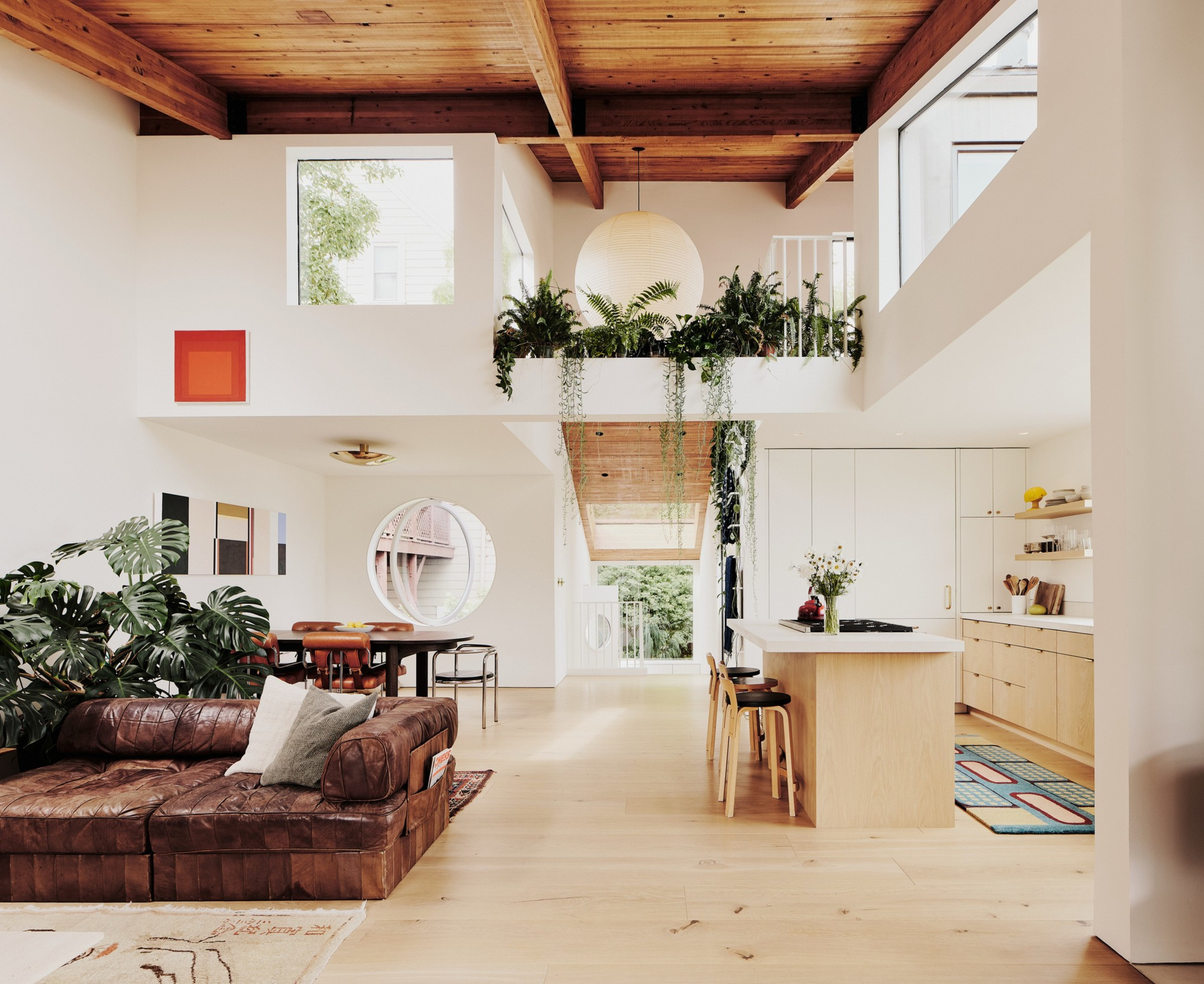 A modern living space with wooden ceiling beams features a kitchen with a central island, a cozy seating area, large plants, and abundant natural light.