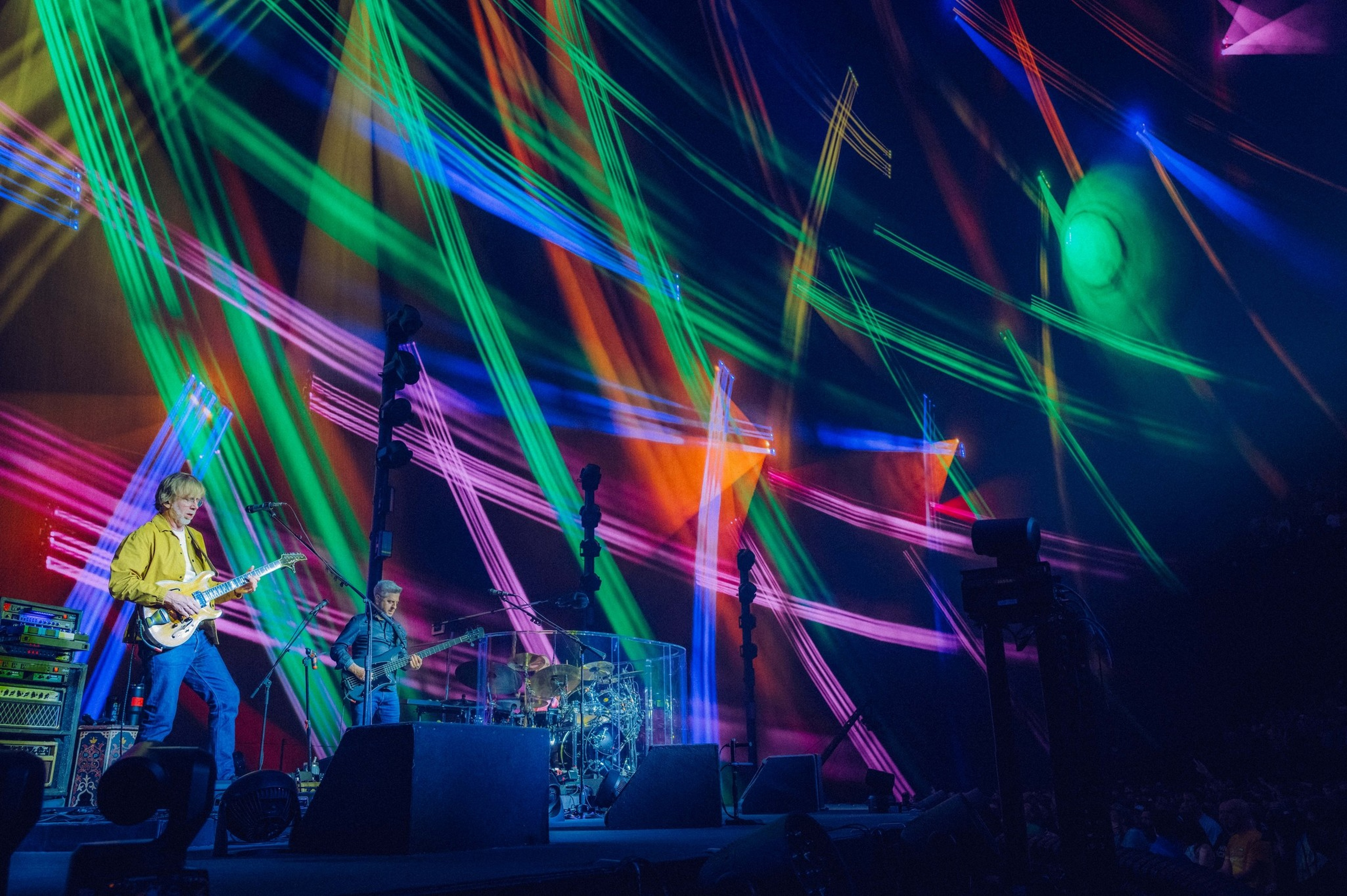 A band performs on stage with vibrant green, blue, pink, and orange laser lights streaming above and around them, creating a dynamic, colorful atmosphere.