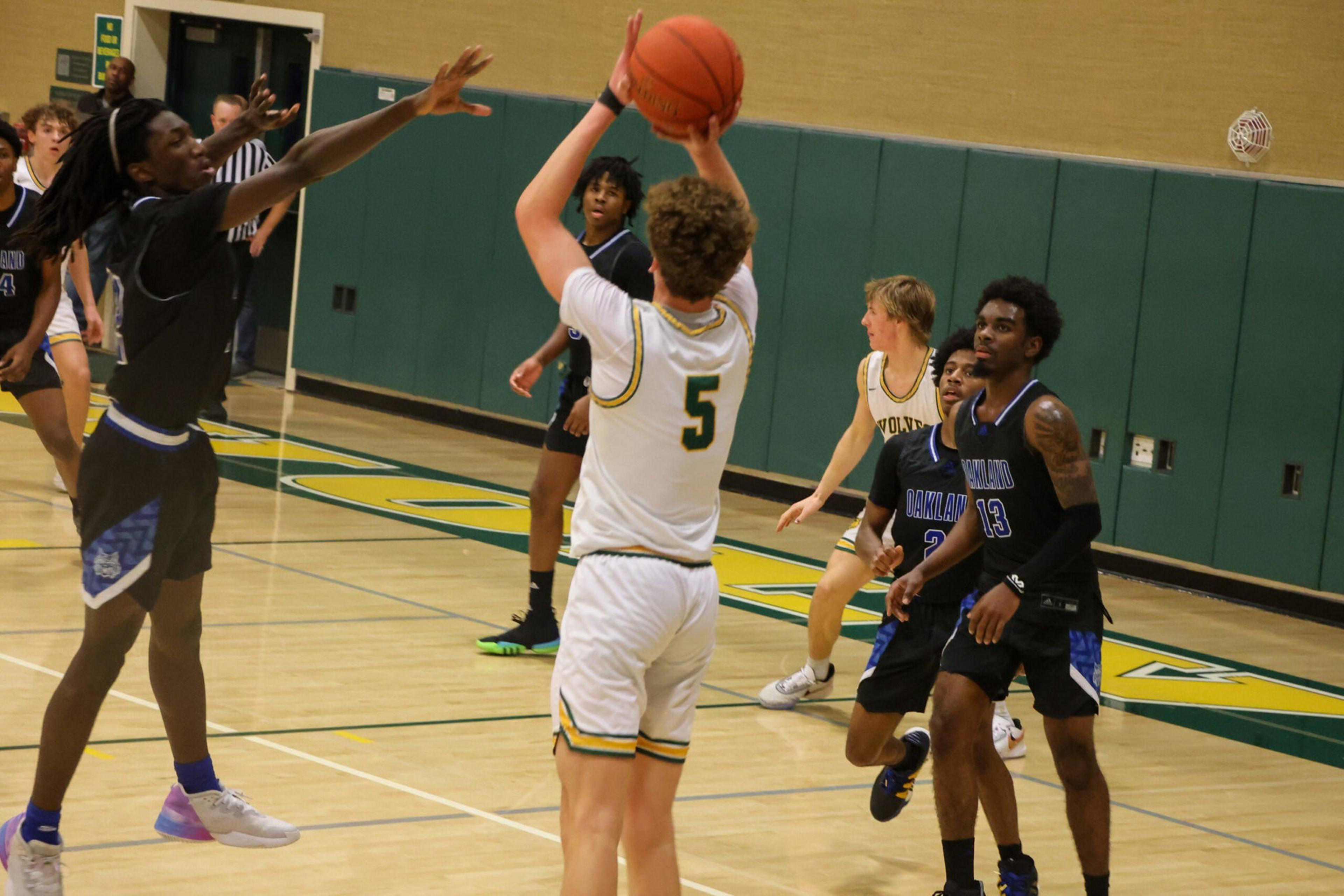 A basketball player in white jersey number 5 jumps to shoot while a defender in black tries to block the shot during a game in an indoor gym.
