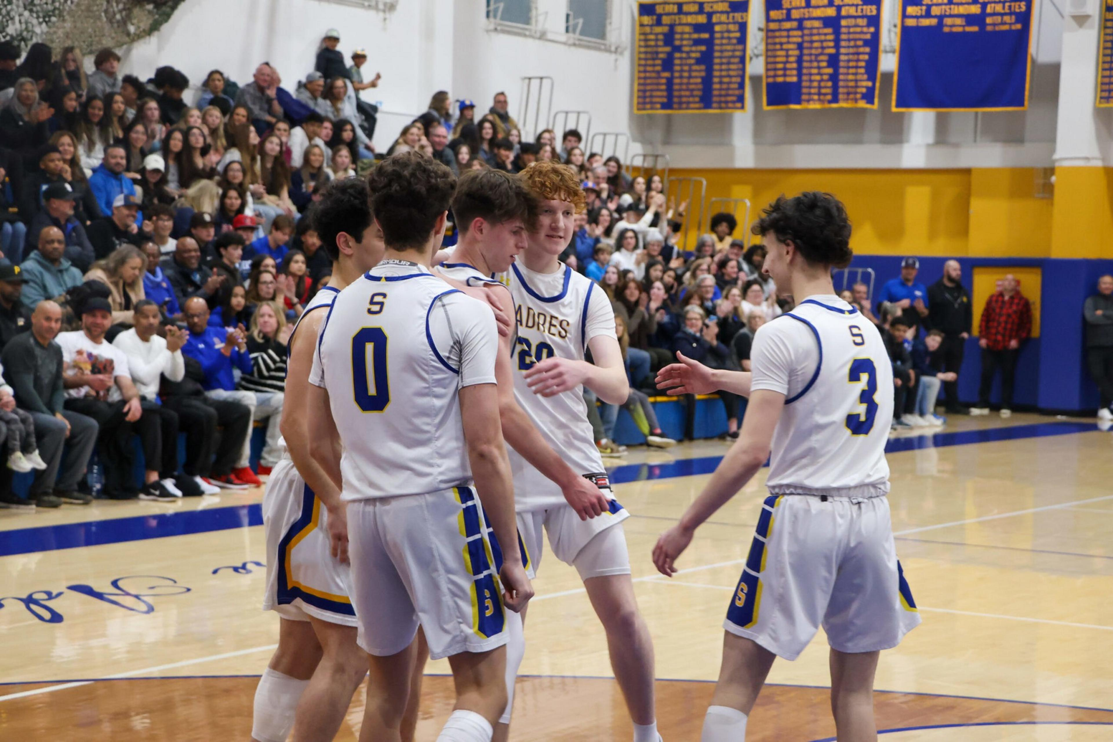 Five basketball players in white and blue uniforms huddle on a court, while a crowd watches from bleachers in a gym with yellow and blue walls.
