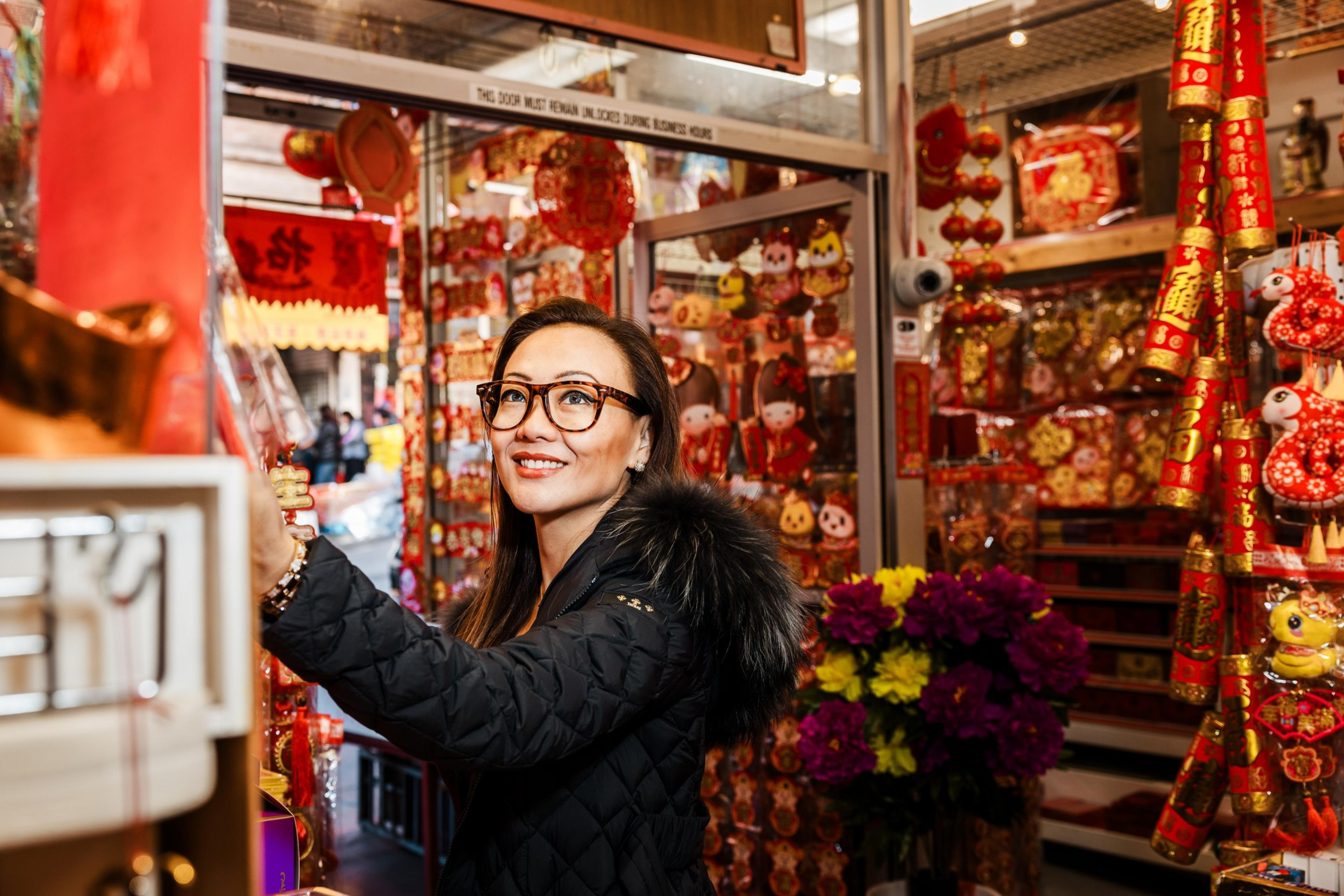 A woman in glasses and a black coat smiles while browsing a colorful shop filled with red and gold decorations, symbolizing a festive atmosphere.