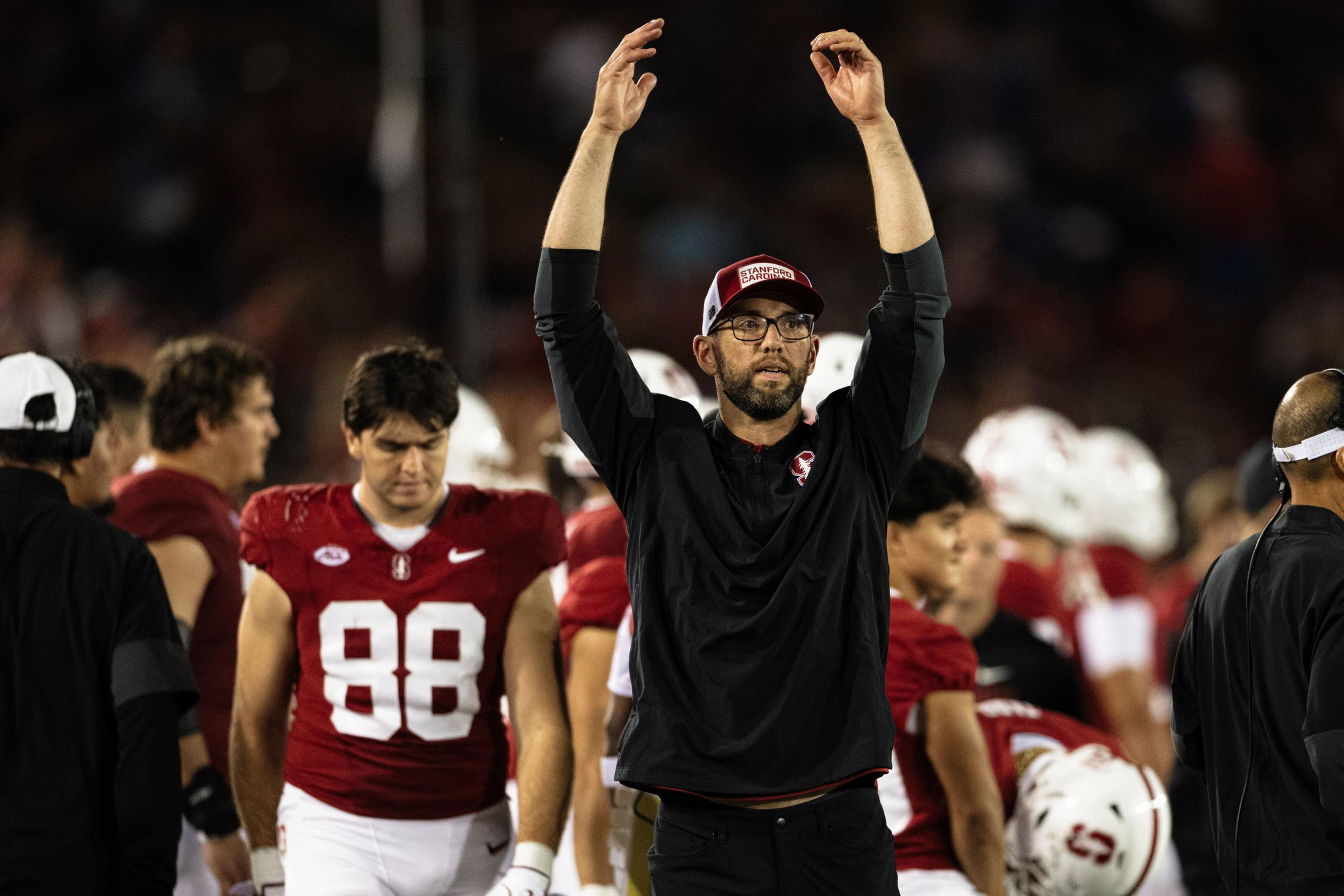 A man wearing glasses, a hat, and black shirt raises both hands, surrounded by football players in red uniforms numbered 88 and others on the field.