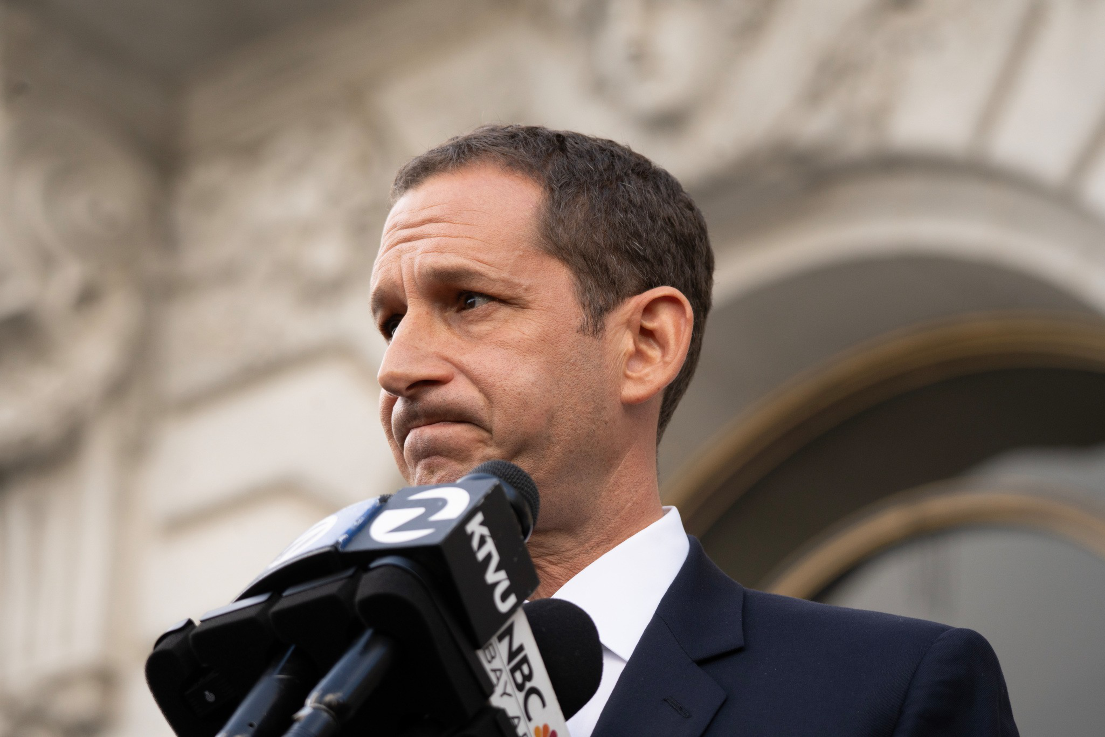 A man in a dark suit and white shirt looks to the side with a serious expression while speaking into multiple microphones outside a stone building.
