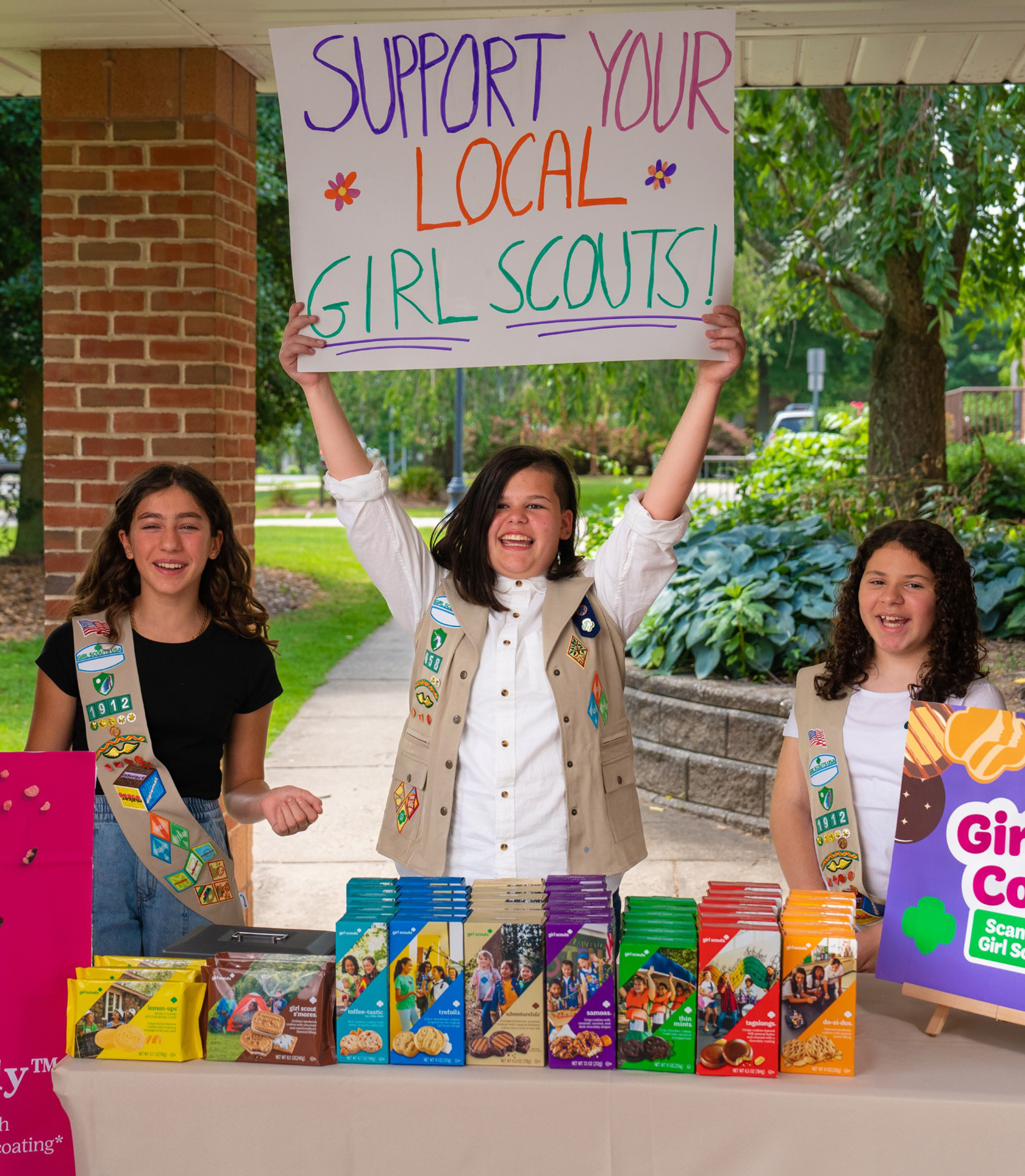 Three Girl Scouts stand behind a table of various Girl Scout cookie boxes, with the middle girl holding a colorful sign that says "Support Your Local Girl Scouts!"