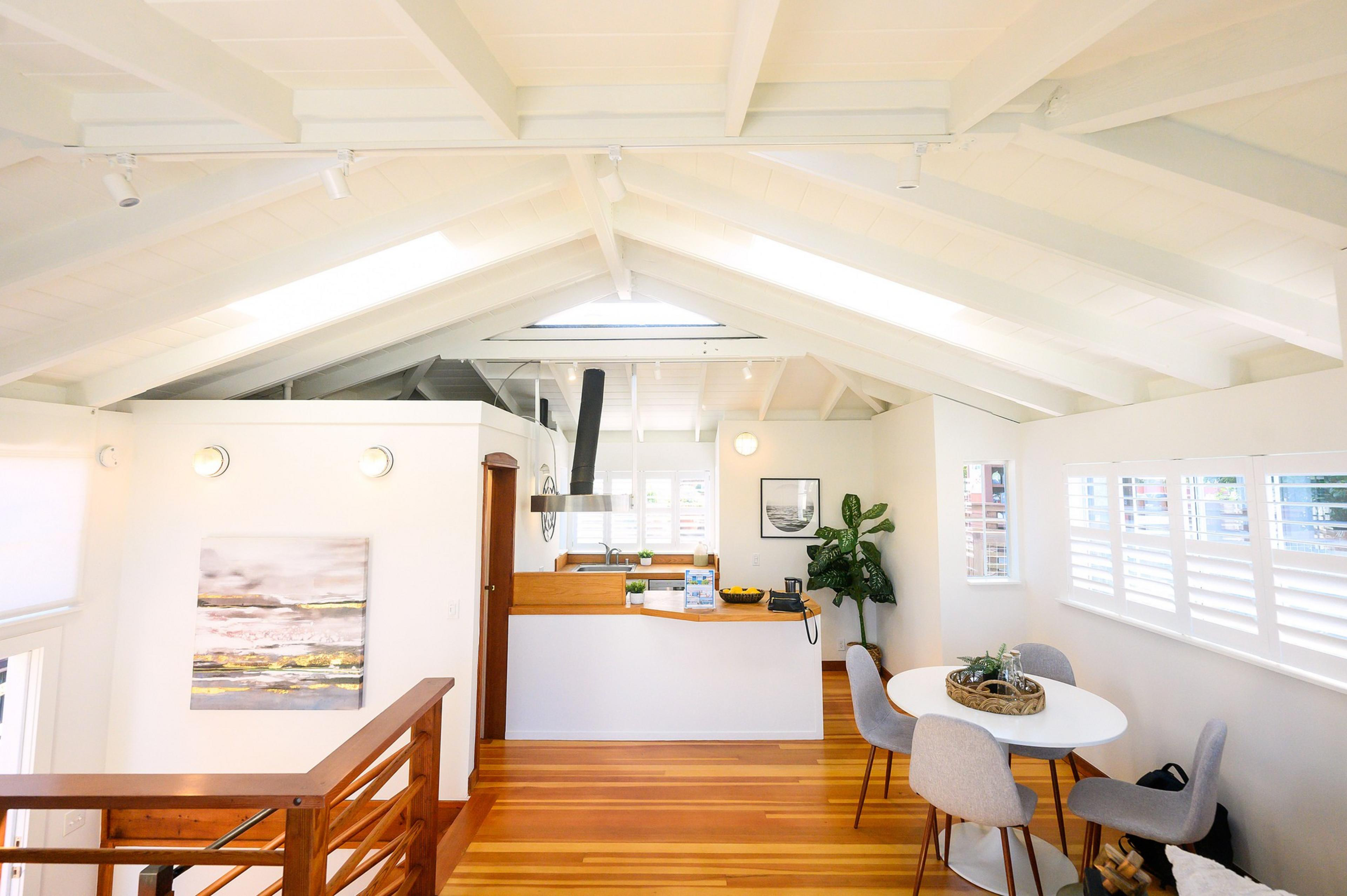 A bright, airy loft space with white walls, wooden floors, a high ceiling, a kitchen area, and a small dining table with chairs.