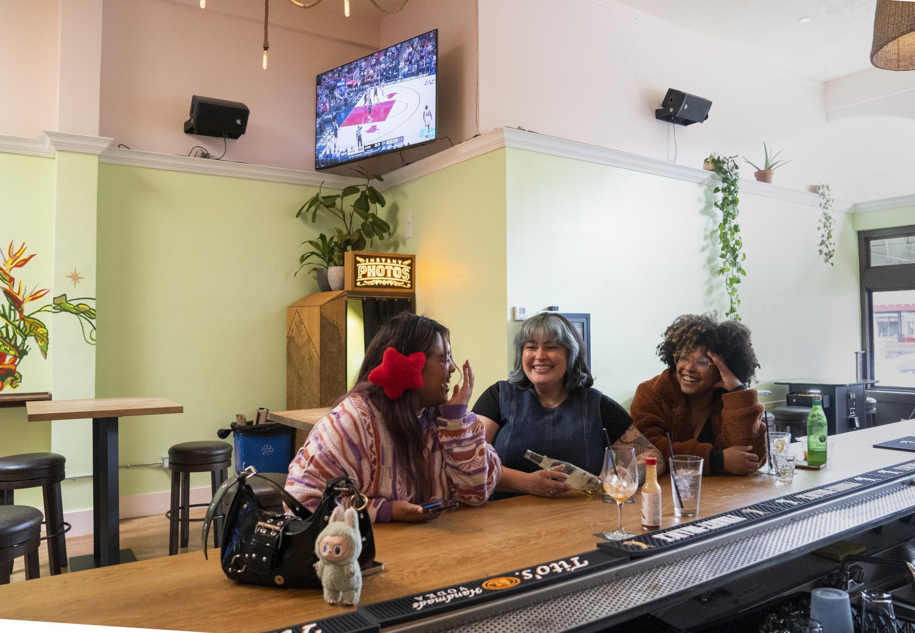 Three people are sitting at a bar, laughing and chatting. A TV above shows a basketball game. There are plants and a photo booth in the background.
