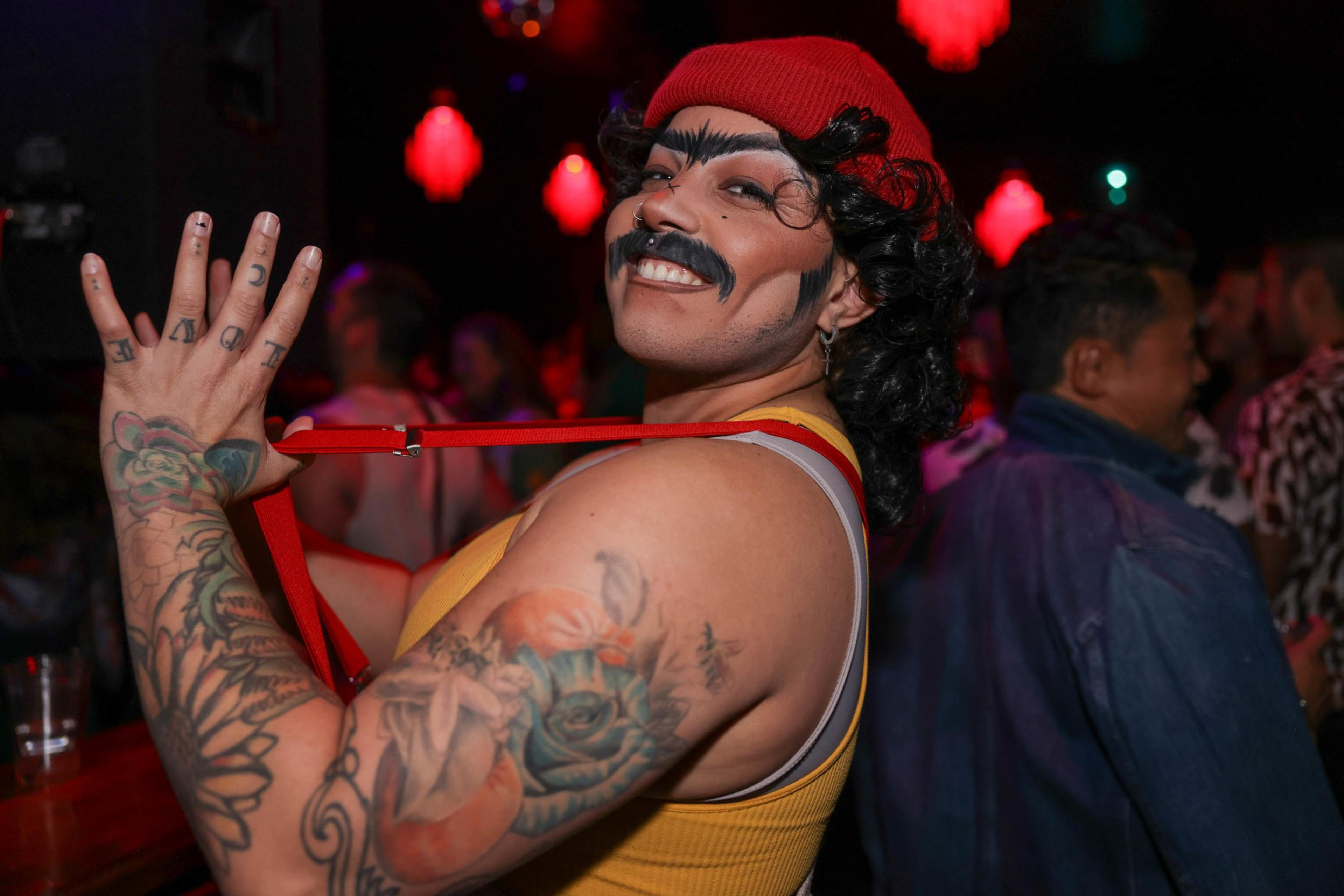 a smiling, mustachioed person in a red hat flicks a red suspender, their tattooed arm exposed