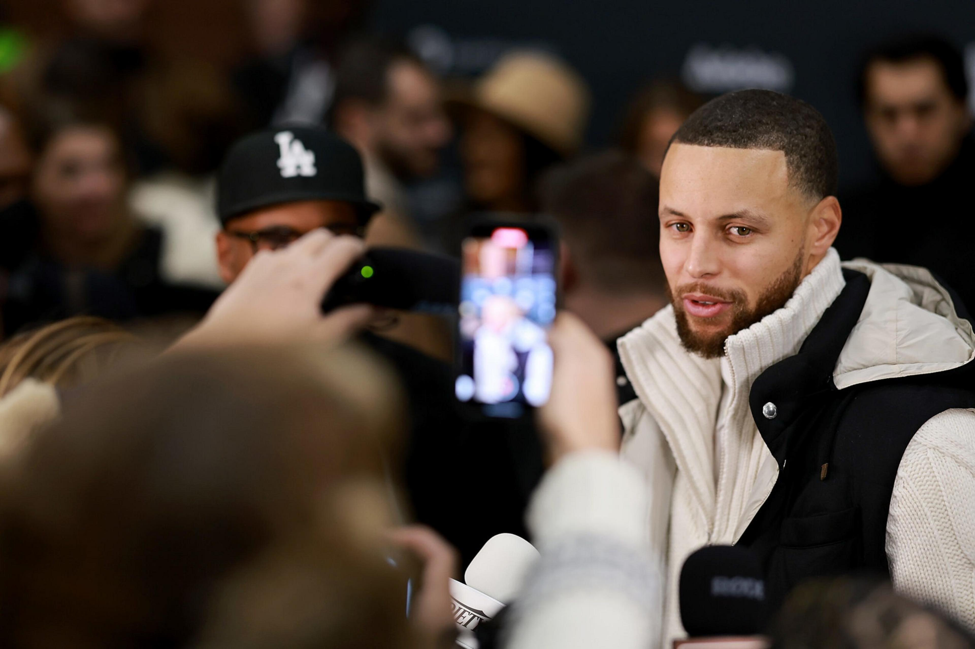 A man with a beard and short hair wears a white turtleneck and black vest while people take photos of him with phones.