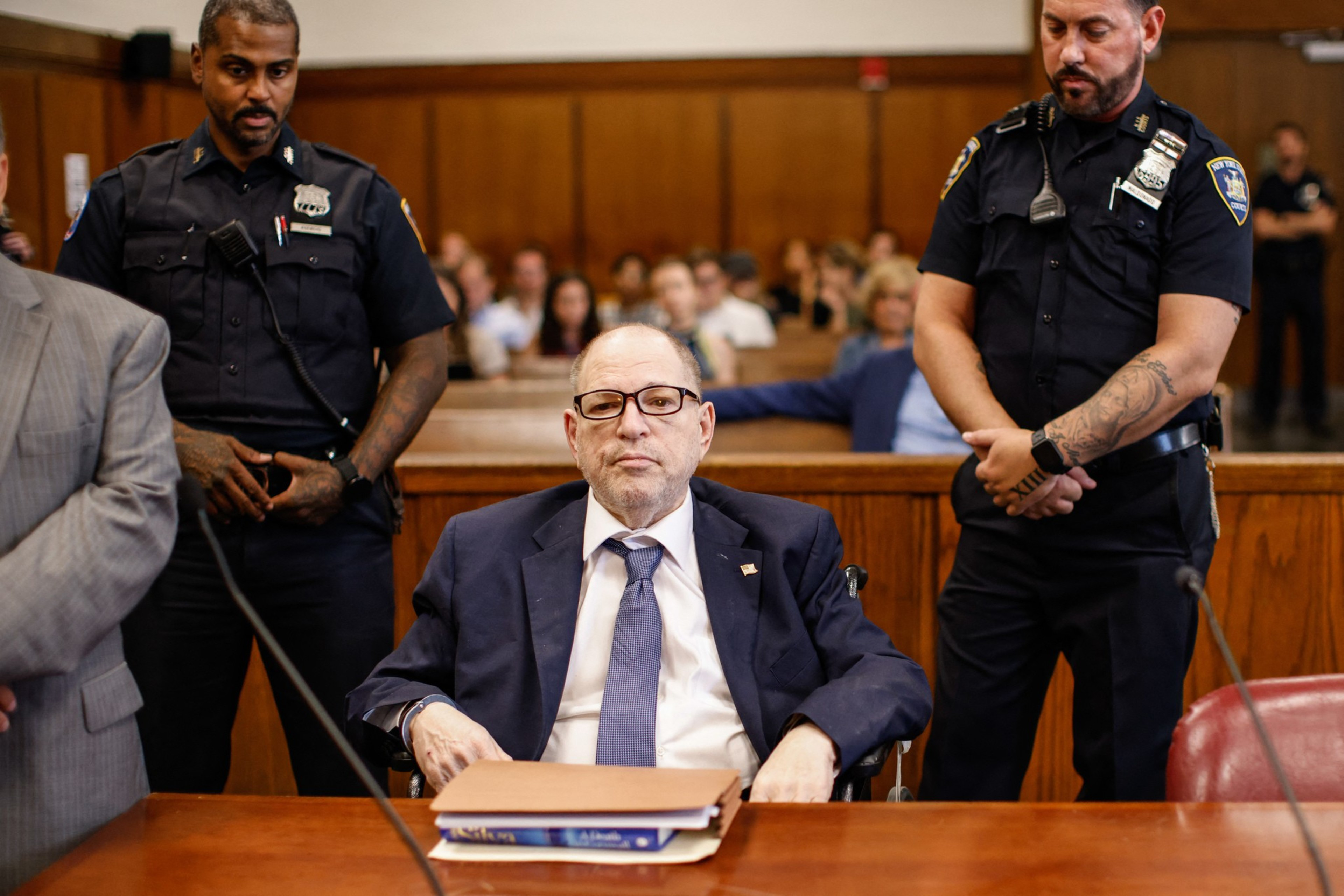 A man in a suit sits at a courtroom table with folders. He's flanked by two uniformed officers, one on each side, while several people are seated in the background.