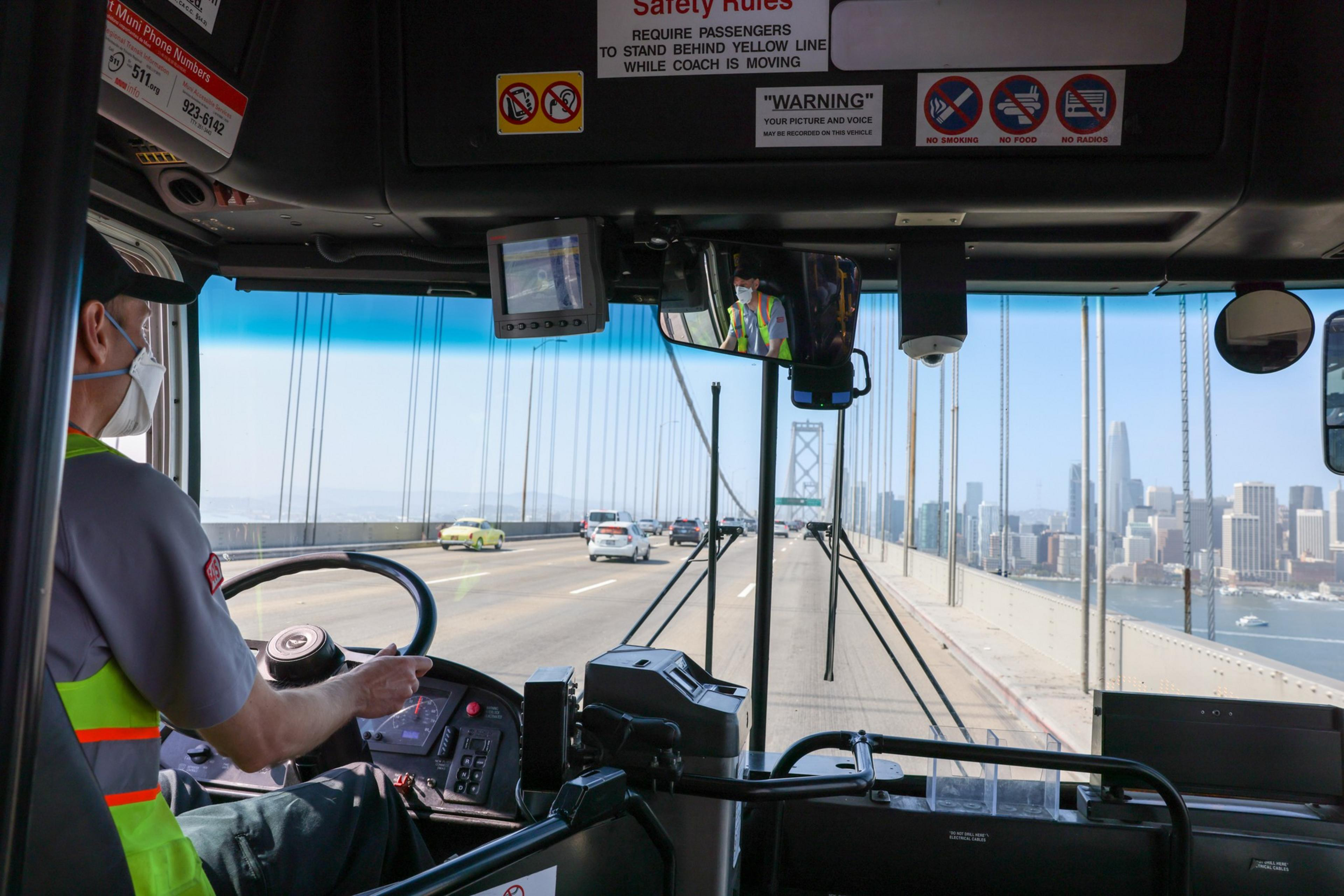 A masked bus driver steers across a bridge with cars on the left lane and a city skyline visible on the right over water.