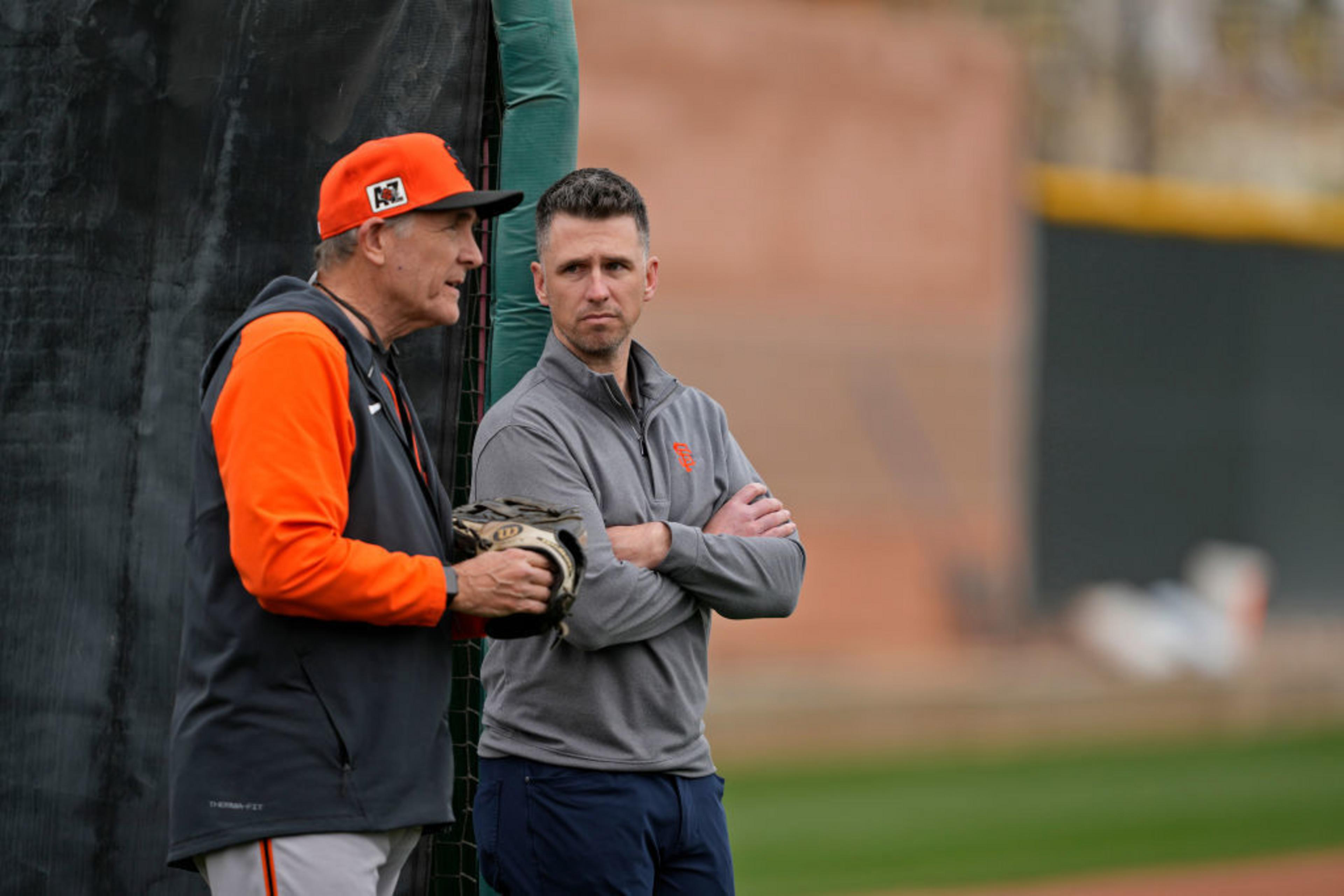 Two men stand on a baseball field. One wears an orange cap and holds a glove, while the other is in a gray jacket with crossed arms. They appear to be in conversation.