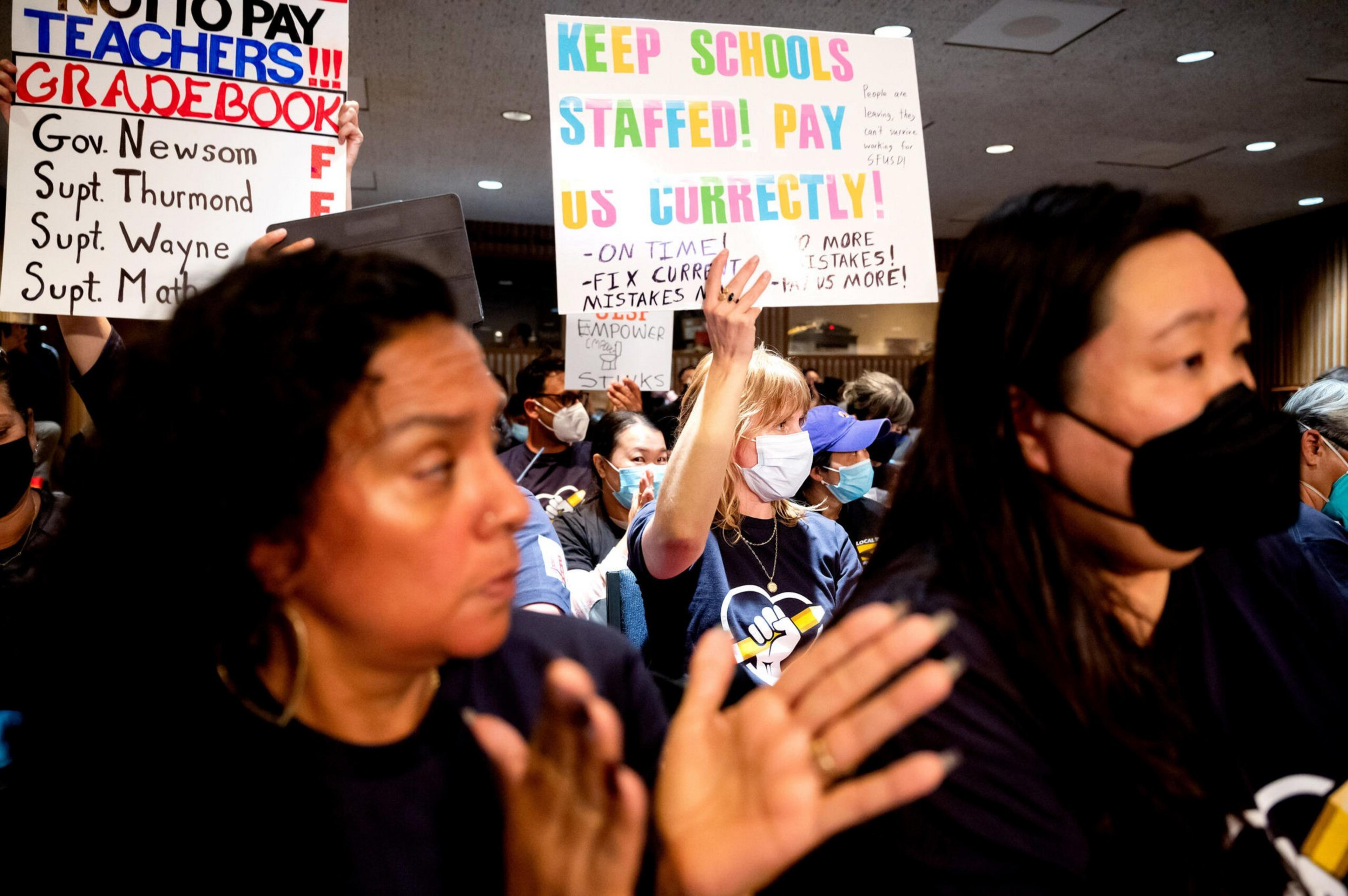 People wearing masks hold colorful protest signs demanding proper pay and staffing for schools, while others clap in support.