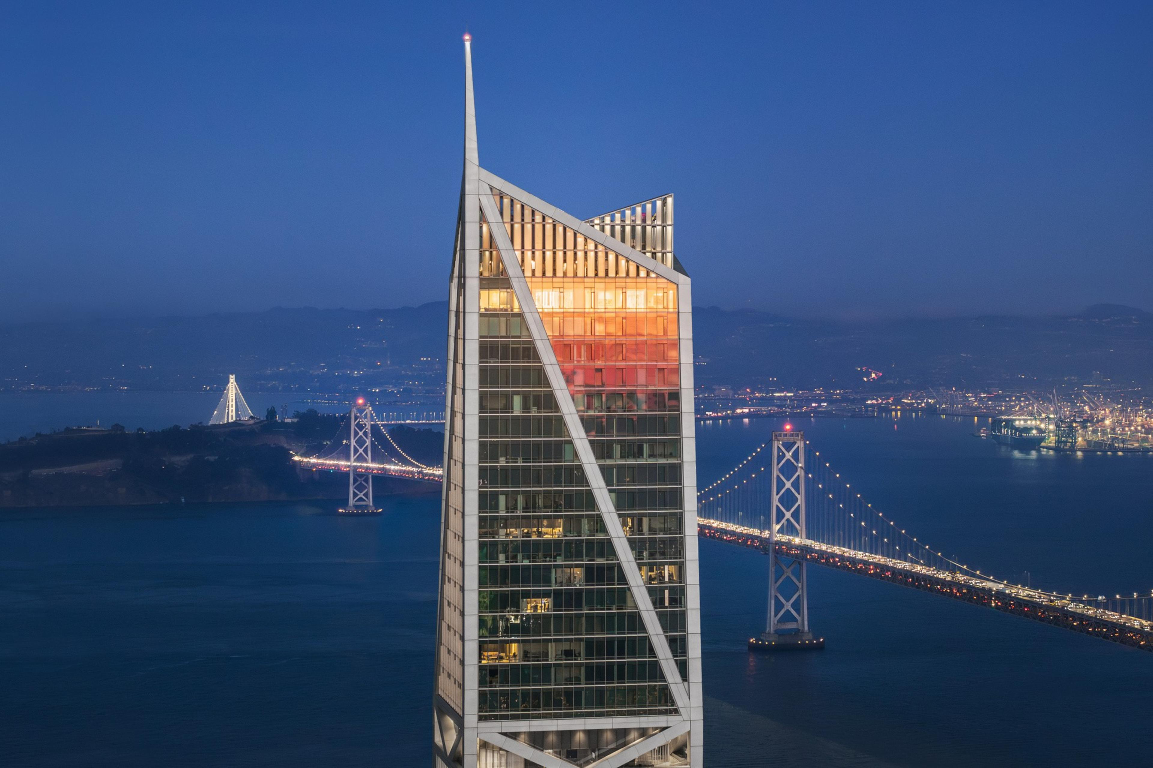 A tall modern skyscraper with a triangular top reflects the sunset, with a lit suspension bridge and city lights in the background over a body of water.