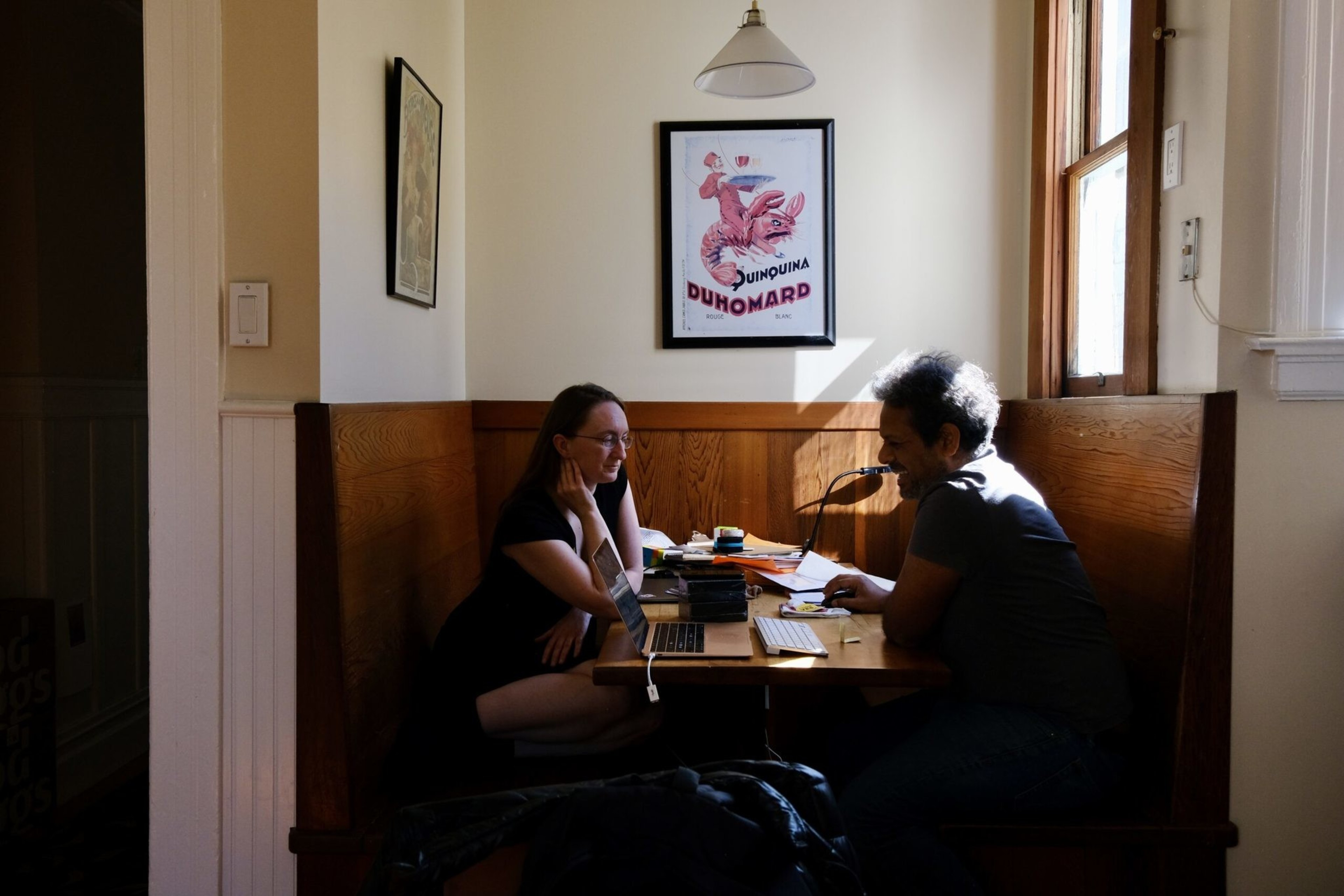 Two people sit across from each other in a wooden booth, working with a laptop, keyboard, and papers, framed by a wall poster and sunlight from a window.