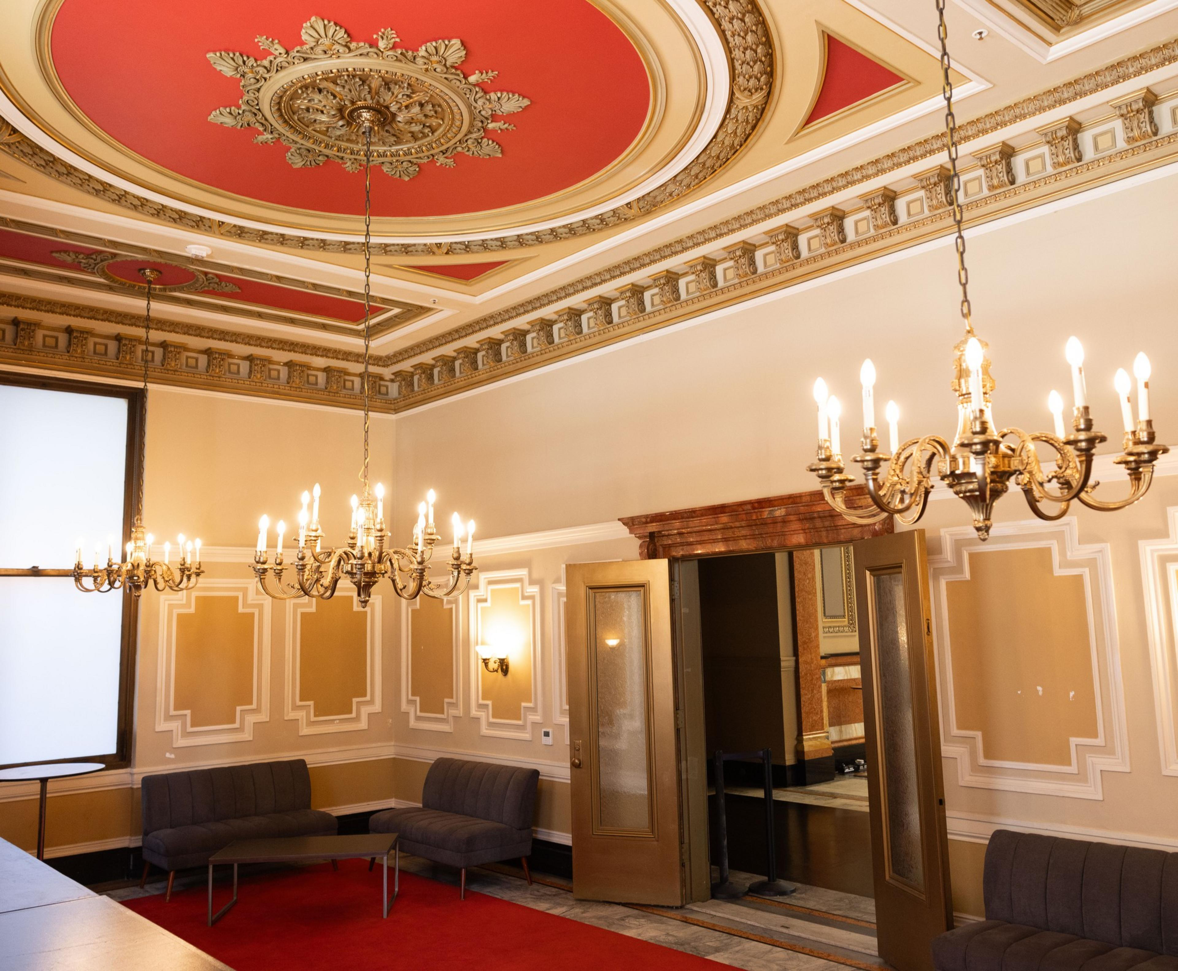 The image depicts an opulent room with ornate, red and gold ceiling details, elaborate chandeliers, gold-framed double doors, and dark gray seating areas on a red carpet.