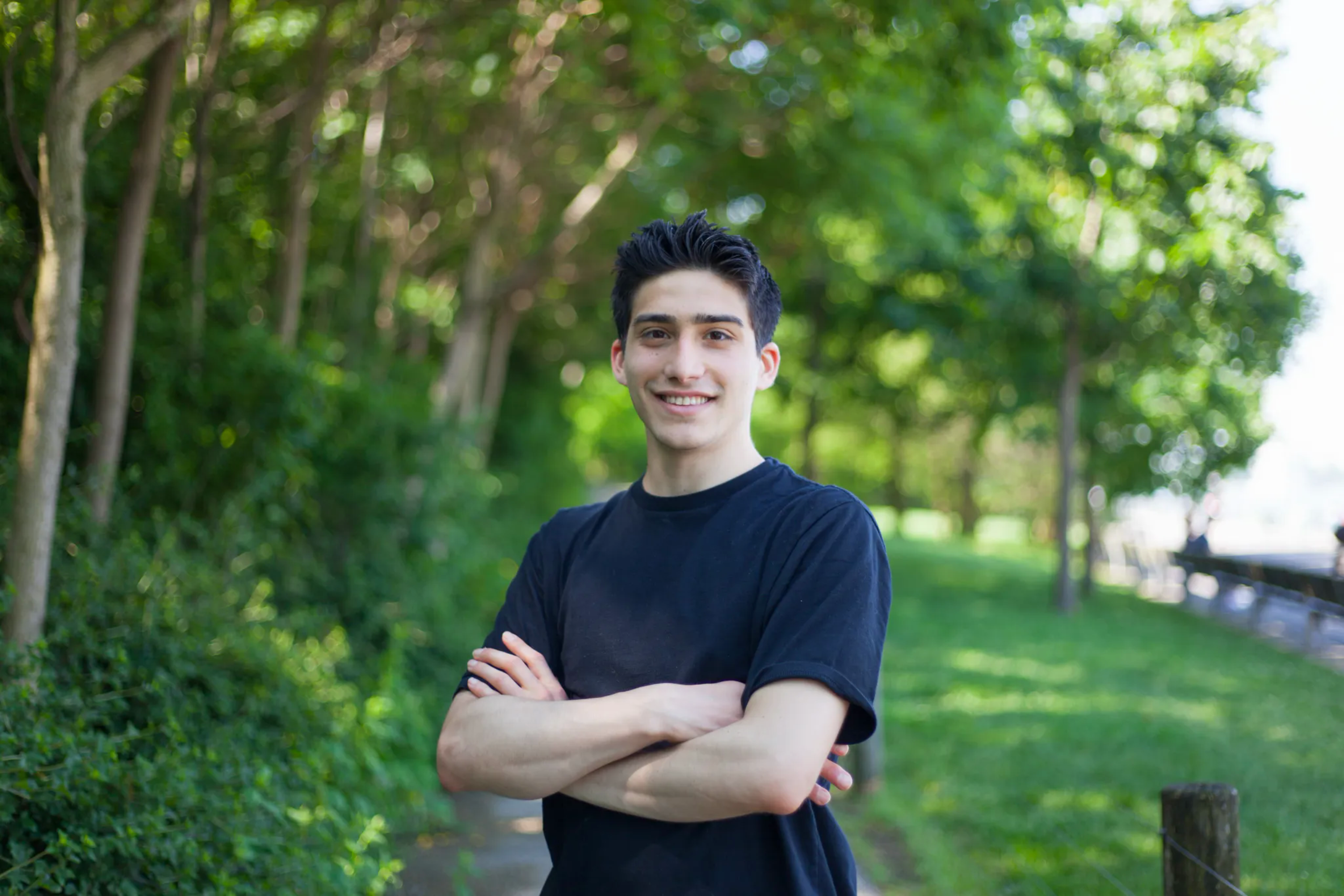 A young man in a black shirt stands outdoors on a tree-lined path, smiling with his arms crossed.