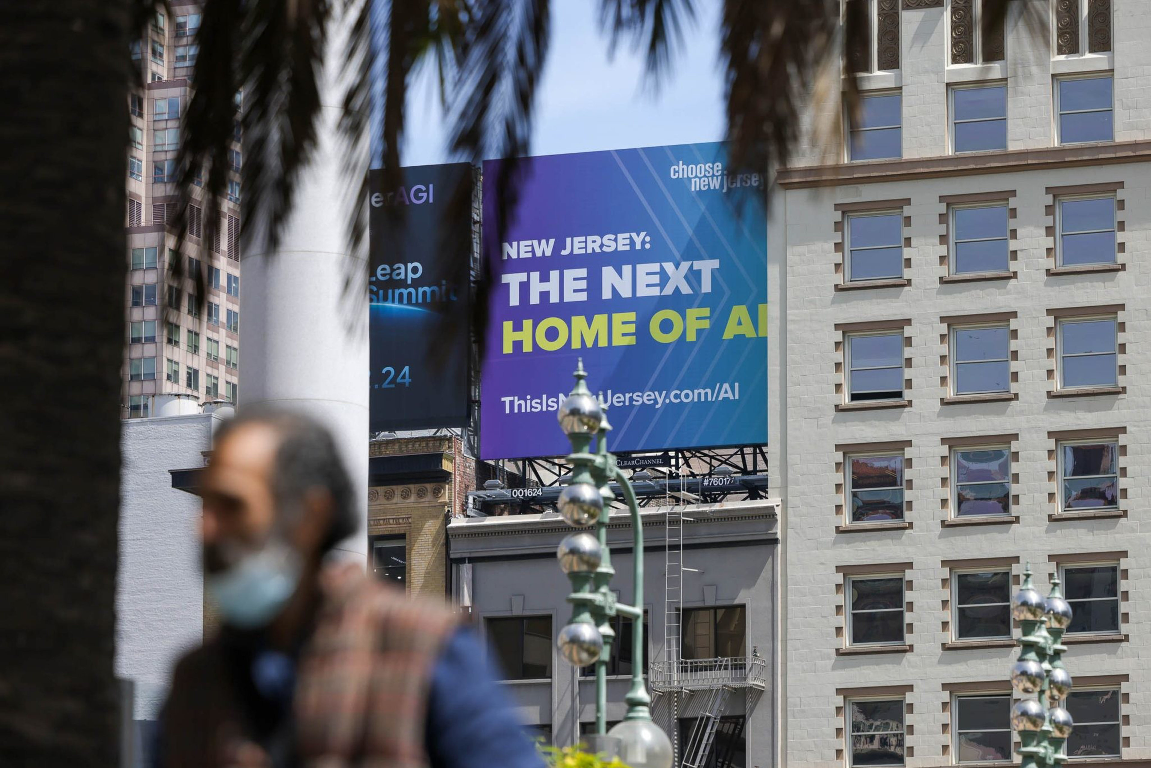 Urban scene with blurred foreground figures, palm trees, a building backdrop, and a billboard promoting New Jersey as "The Next Home of AI."