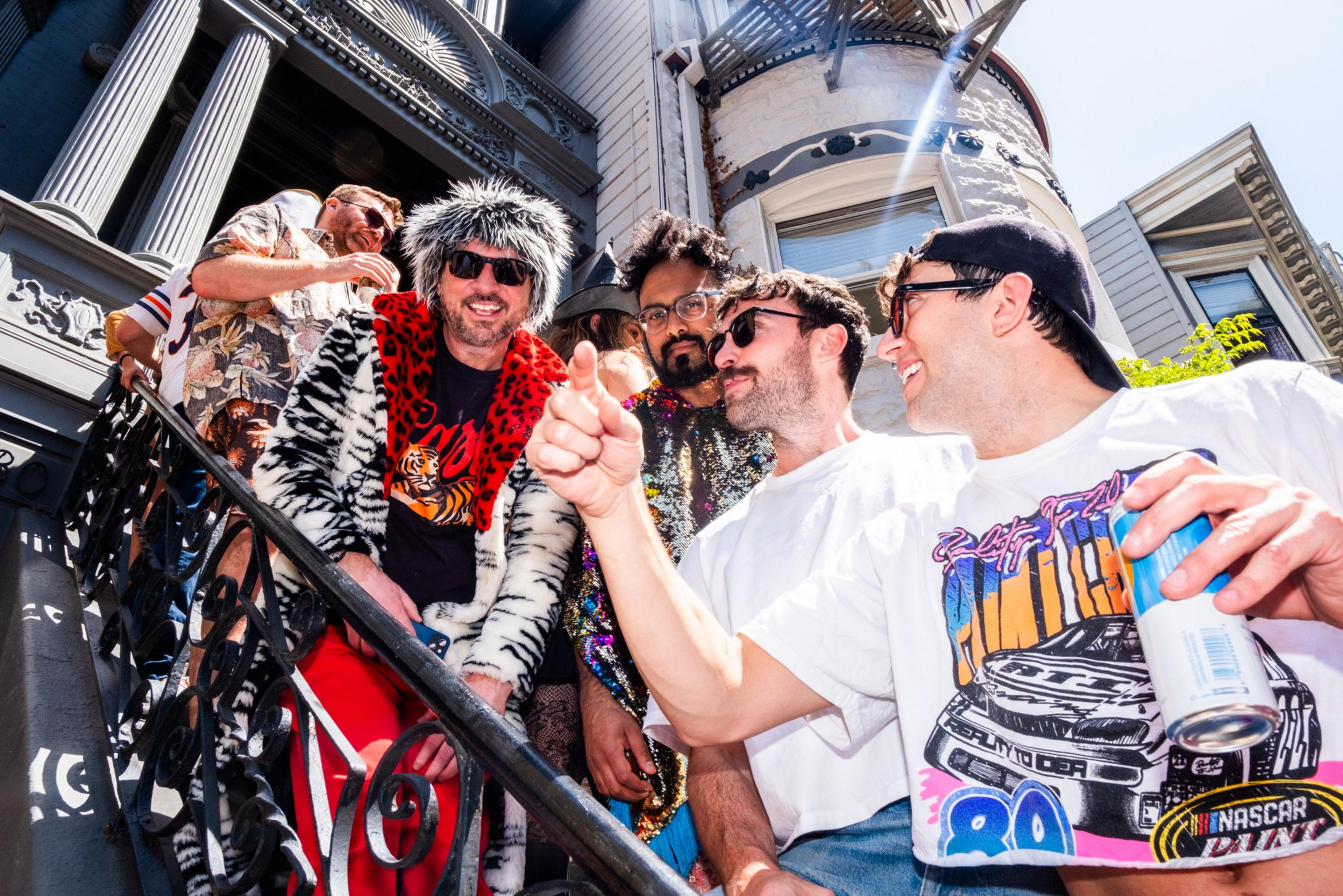 A group of five people stands on a staircase outside a building. They wear colorful, eccentric outfits, including fur and sequin tops, and are smiling and chatting.