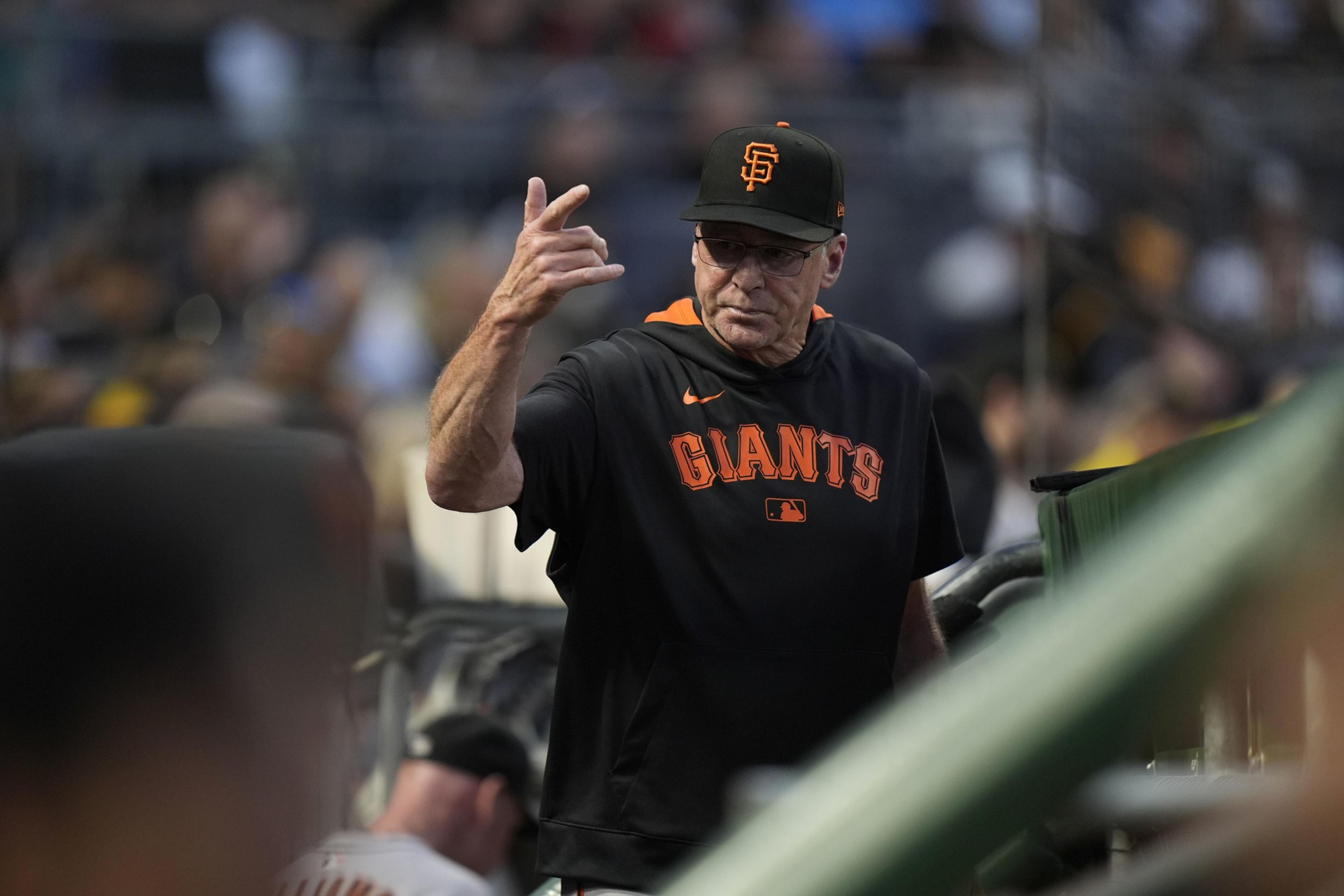 A man wearing a black San Francisco Giants shirt and cap gestures with his hand while standing near a dugout in a baseball stadium.