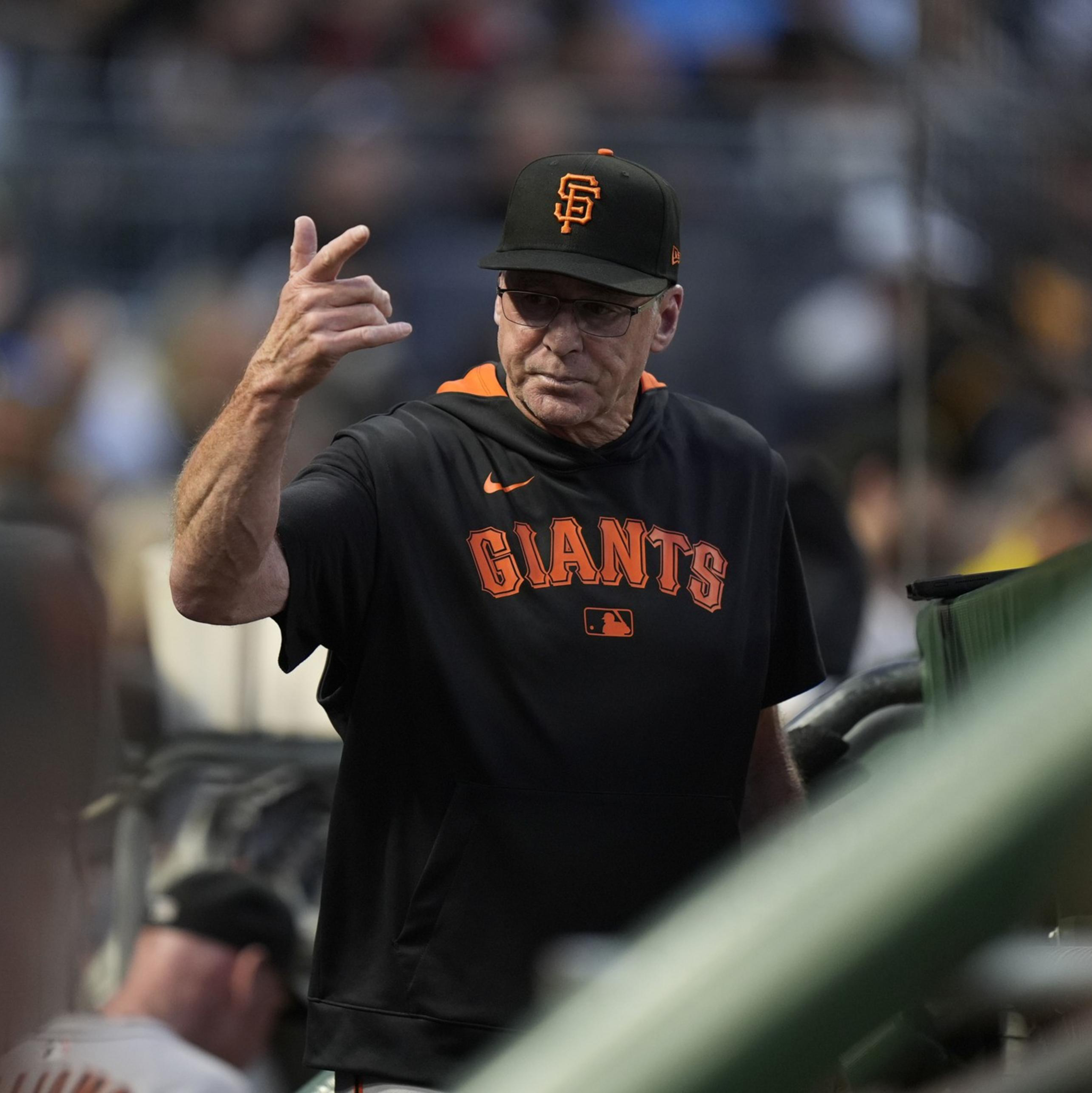 A man wearing a black San Francisco Giants shirt and cap gestures with his hand while standing near a dugout in a baseball stadium.