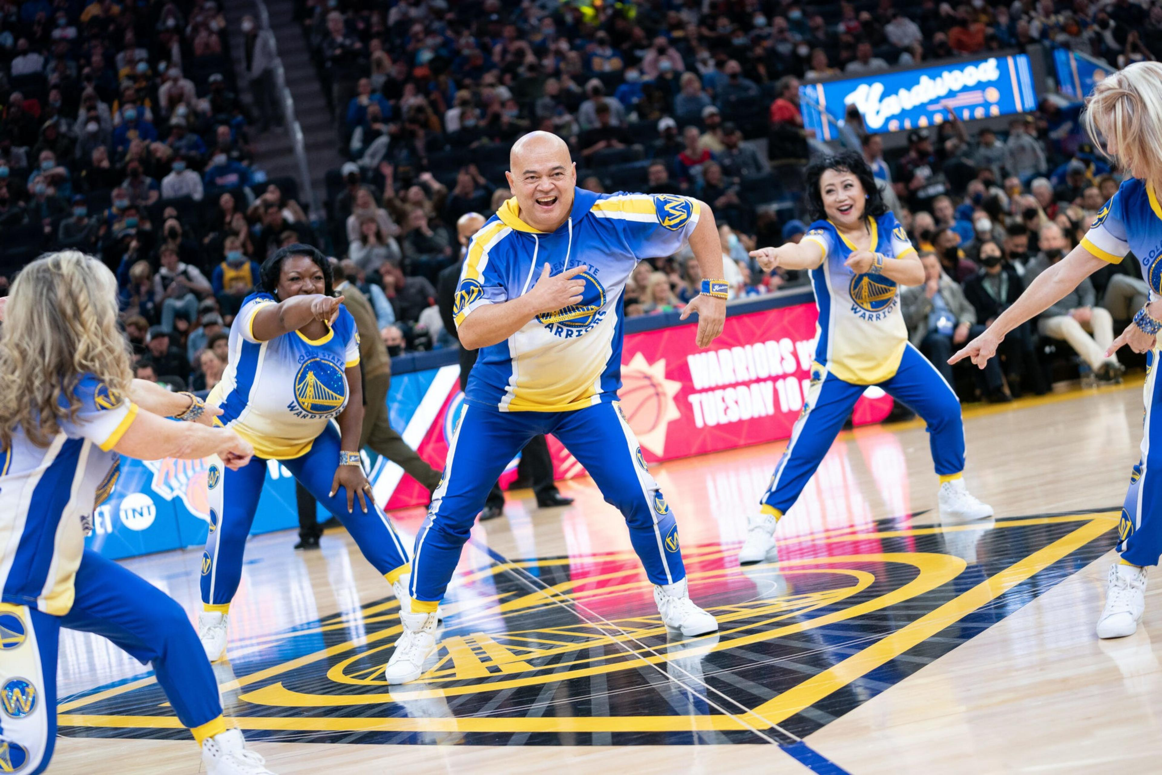 Five people wearing Golden State Warriors dance uniforms perform a lively routine on a basketball court with an audience in the background.