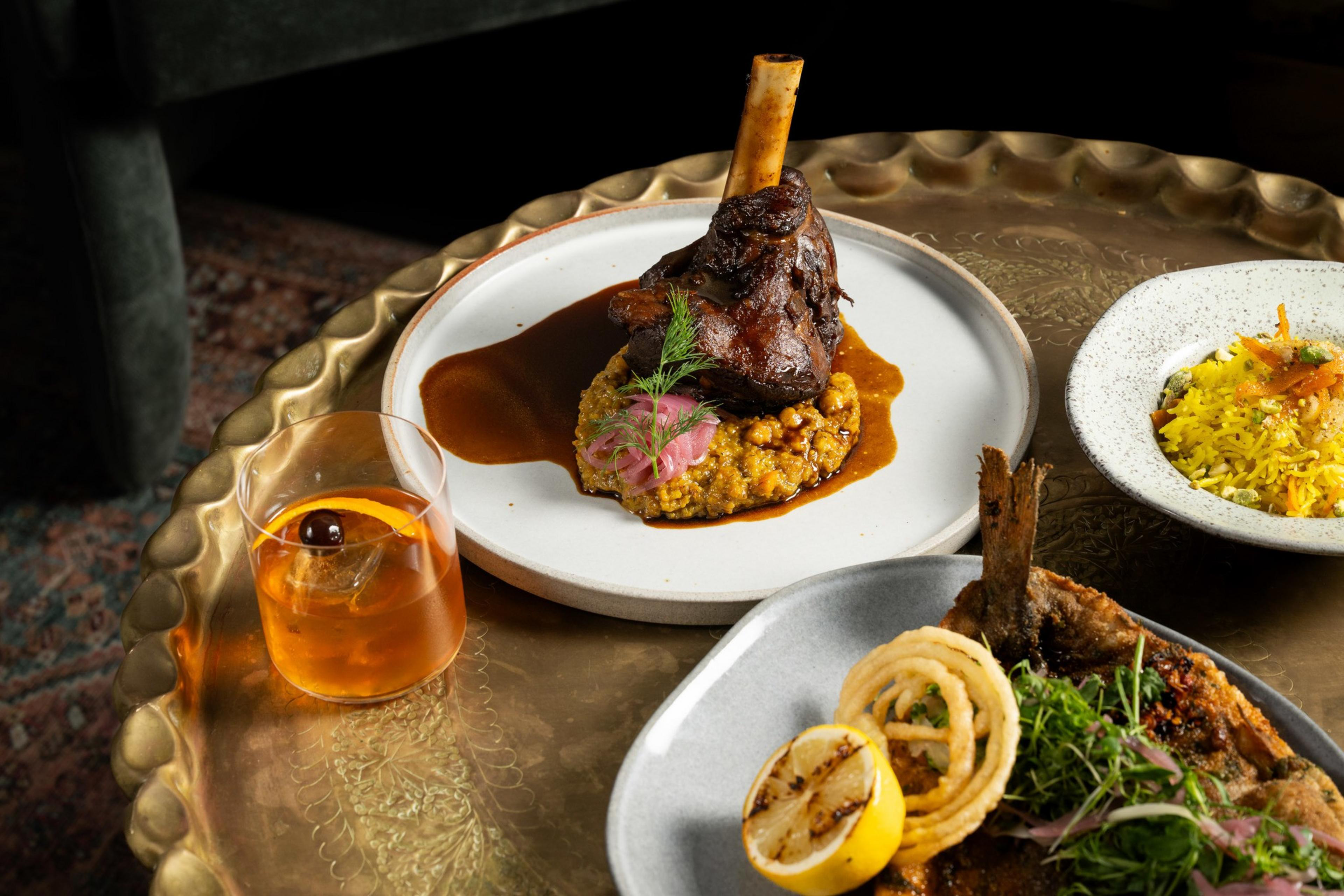 A table features a braised lamb shank on a white plate, turmeric rice in a bowl, and fried fish with lemon and onion rings. A cocktail rests beside the dishes.