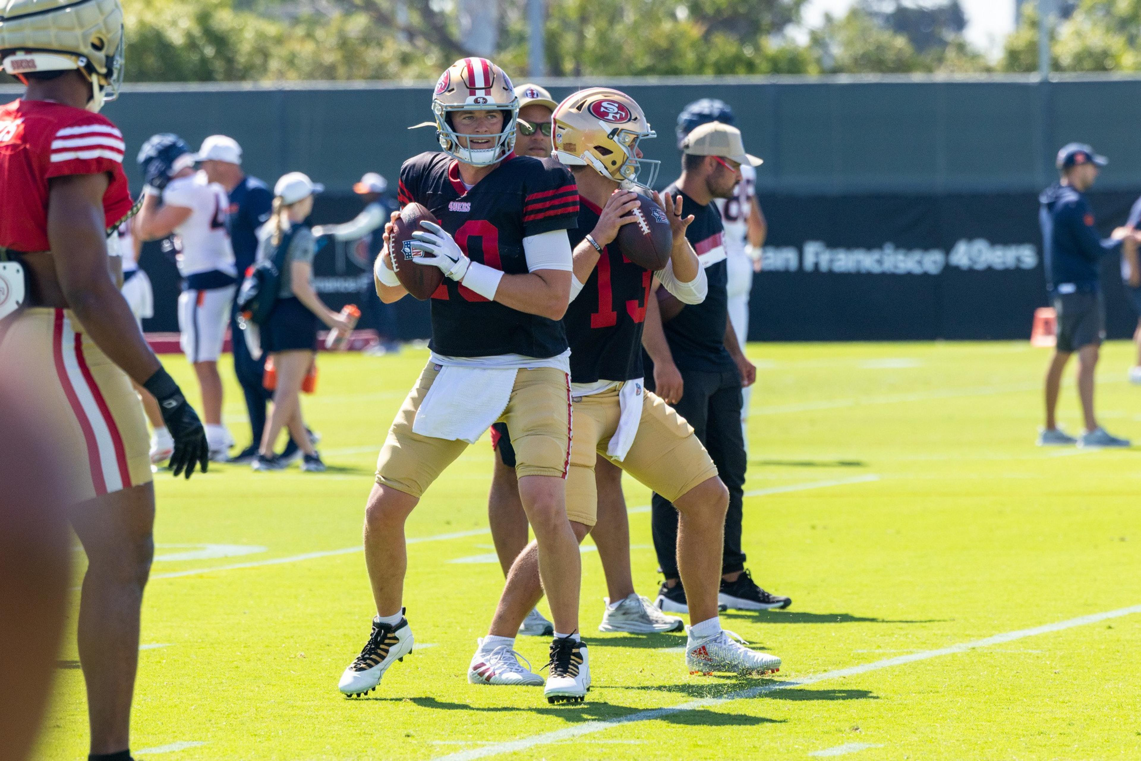 Two football players wearing black jerseys and gold pants practice throwing on a sunny field with others in the background.