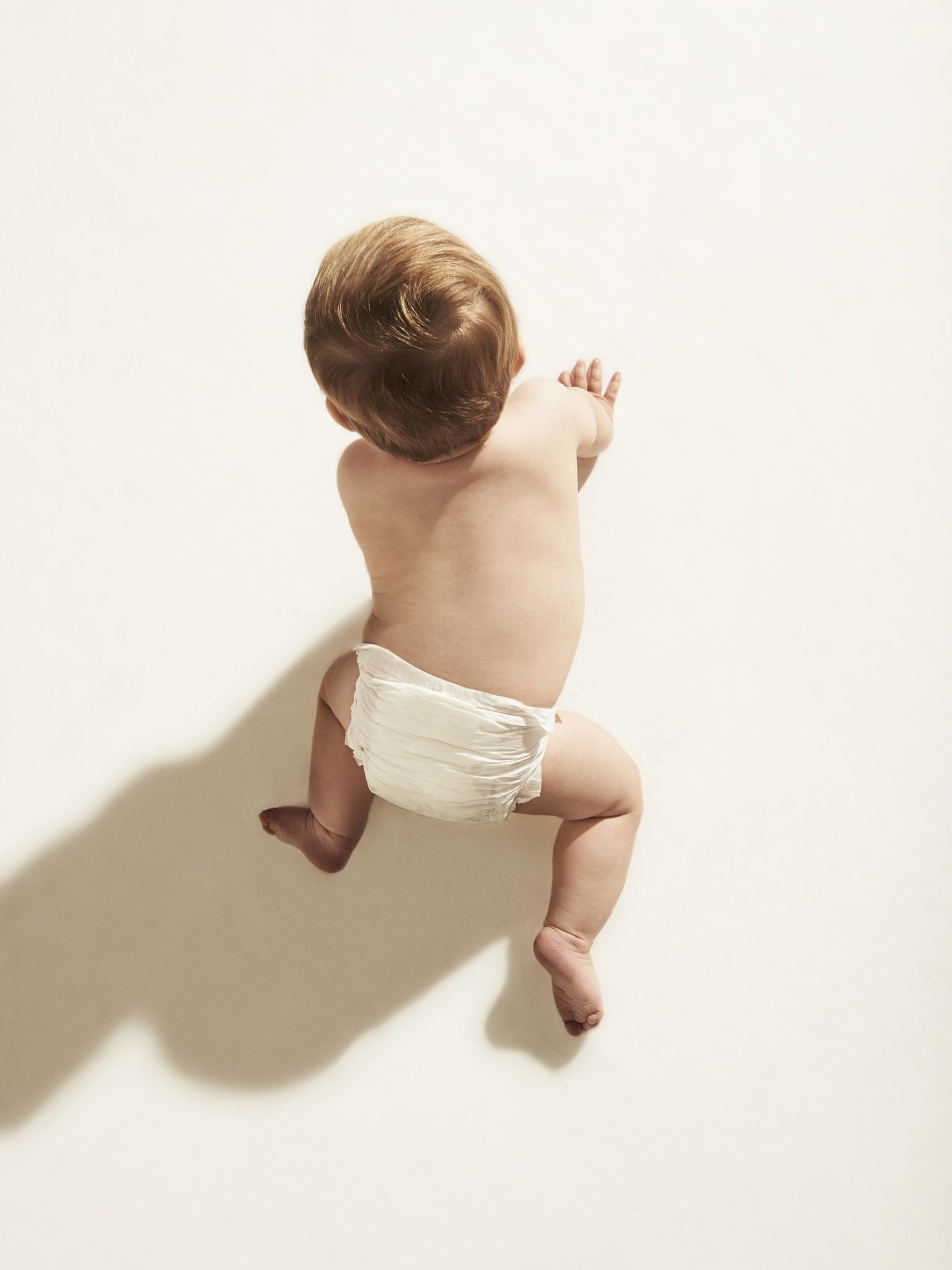A baby in a white diaper is crawling on a light surface, casting a shadow to the left. The image is taken from above, showing the baby's back and soft hair.