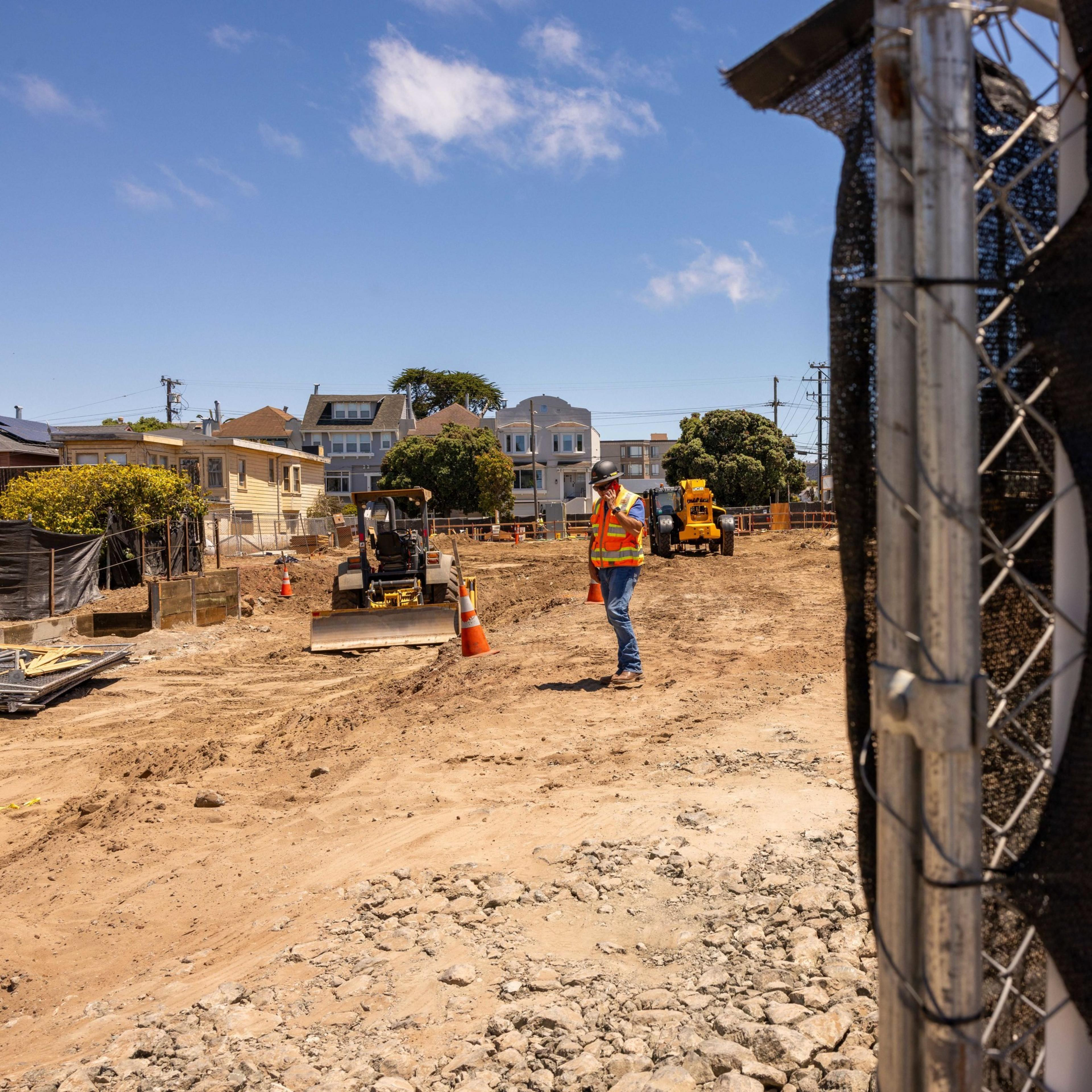 A construction worker in a reflective vest and hard hat stands on a dirt construction site with a small bulldozer and houses in the background, under a clear blue sky.